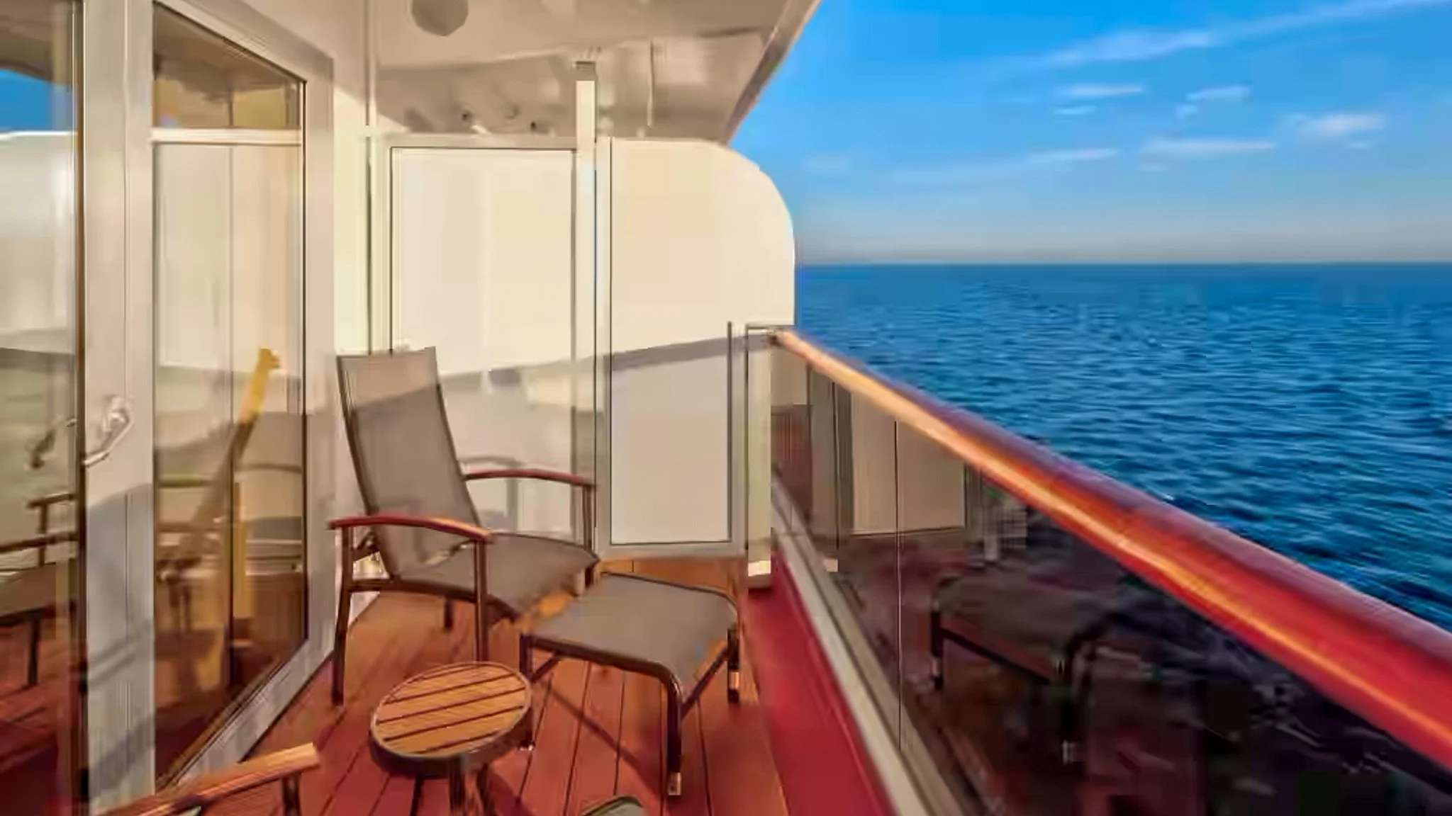 View of a balcony on a cruise ship with chairs and a small table overlooking the ocean under a partly cloudy sky.