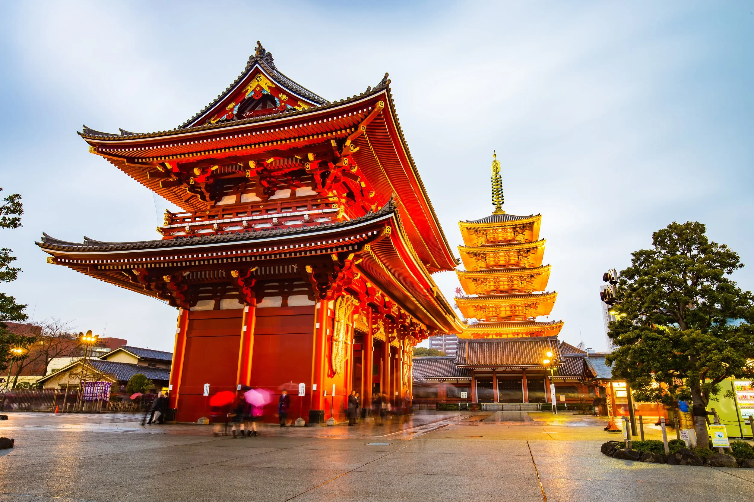 Traditional Japanese temple with red and gold pagoda-style buildings, surrounded by trees and evening sky.
