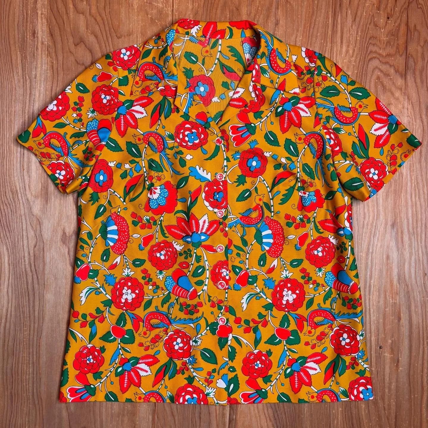 Like a piece fabric torn from a storybook quilt, this vintage button-up blooms with folkloric charm. A kaleidoscope of blooming flowers, twisting vines, and folkloric birds dance across a mustard yellow canvas in a print that feels straight out of an