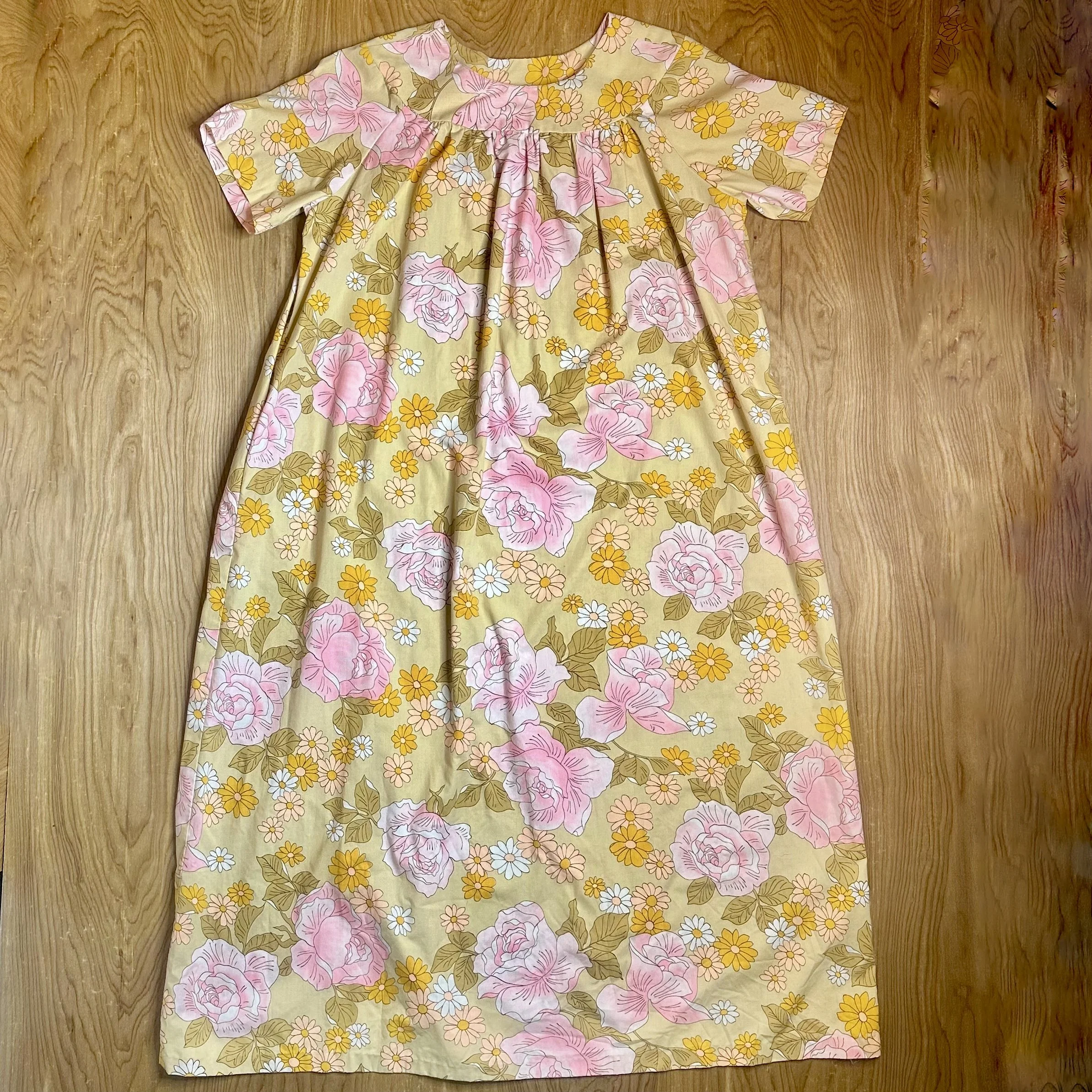 Vintage floral muumuu dress with soft pink roses and yellow daisies, laid flat on wooden floor.