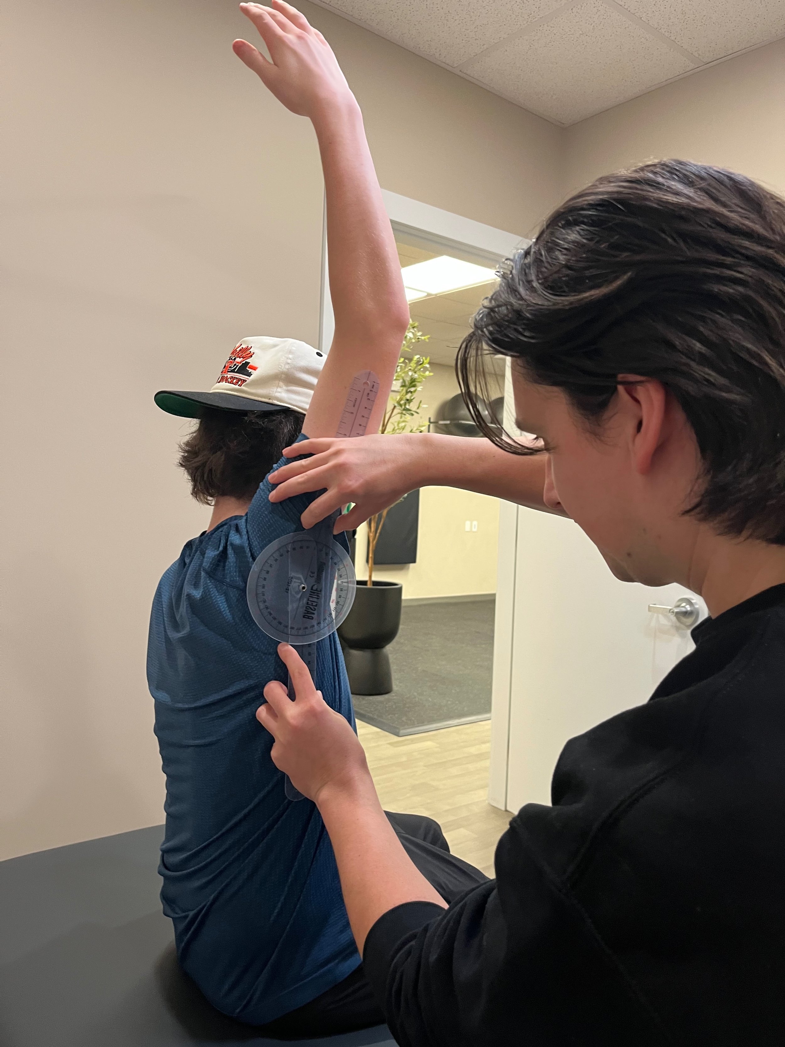 A person receiving a massage on their leg outdoors on a sunny day, with another person providing the massage. The person on the ground is lying on a blue mat with hands on their stomach and eyes closed.