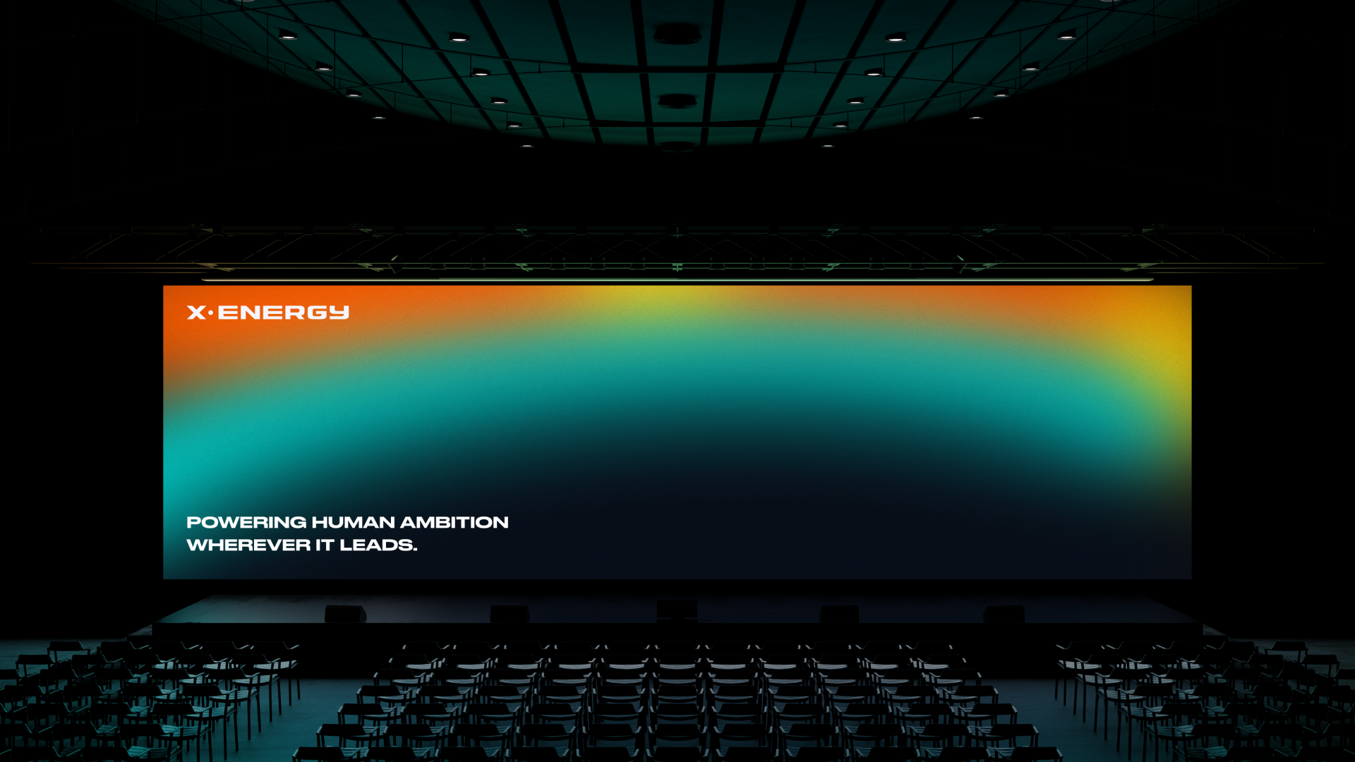 Large presentation screen in a dark auditorium featuring a gradient background with the text 'X-ENERGY' in the top left corner and a slogan underneath that reads 'Powering human ambition wherever it leads.' Rows of empty chairs are arranged facing th