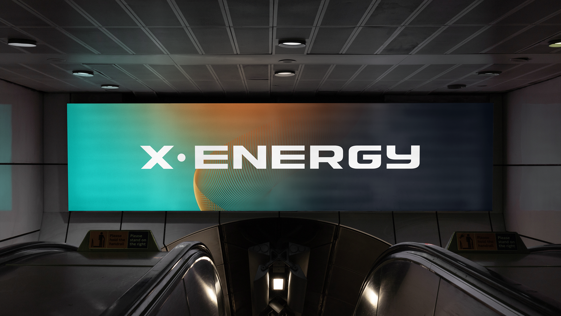 Large illuminated digital sign displaying the word 'X·ENERGY' with a colorful, abstract background, located on an indoor wall above escalators.