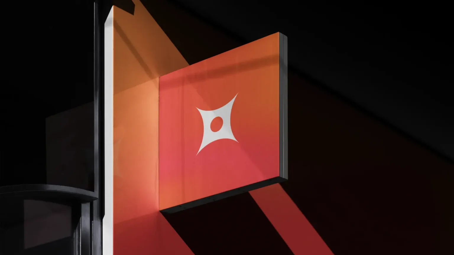 A display screen with a red background and a white abstract logo in the center, mounted on a dark wall.