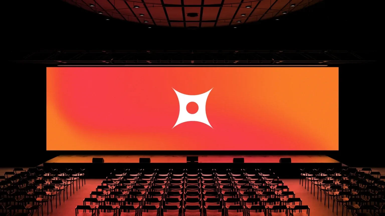 Large auditorium with empty seating facing a brightly lit orange screen with a white logo in the center.