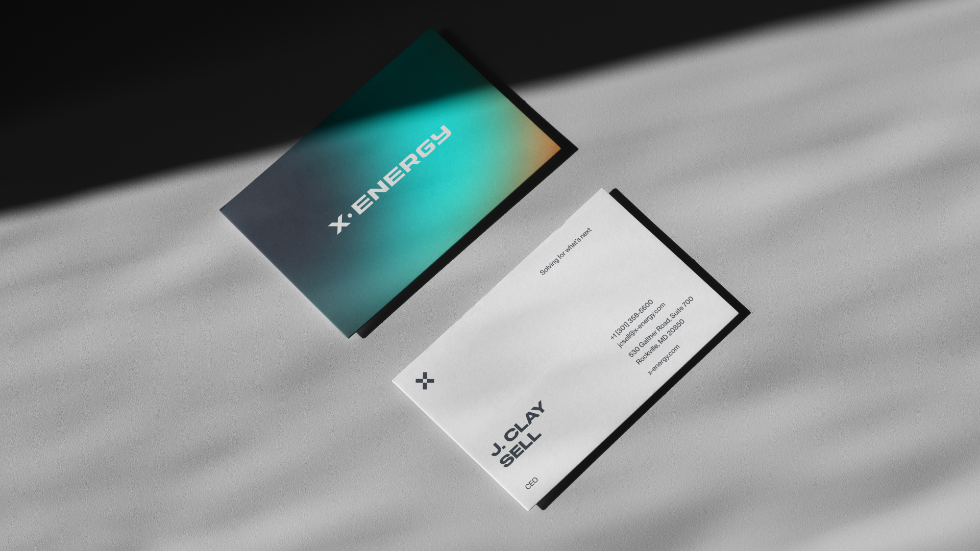 Business card with the logo 'X:ENERGY' on a gradient background and contact information for J. Clay Sell on a white background, both cards casting shadows on a textured grey surface.