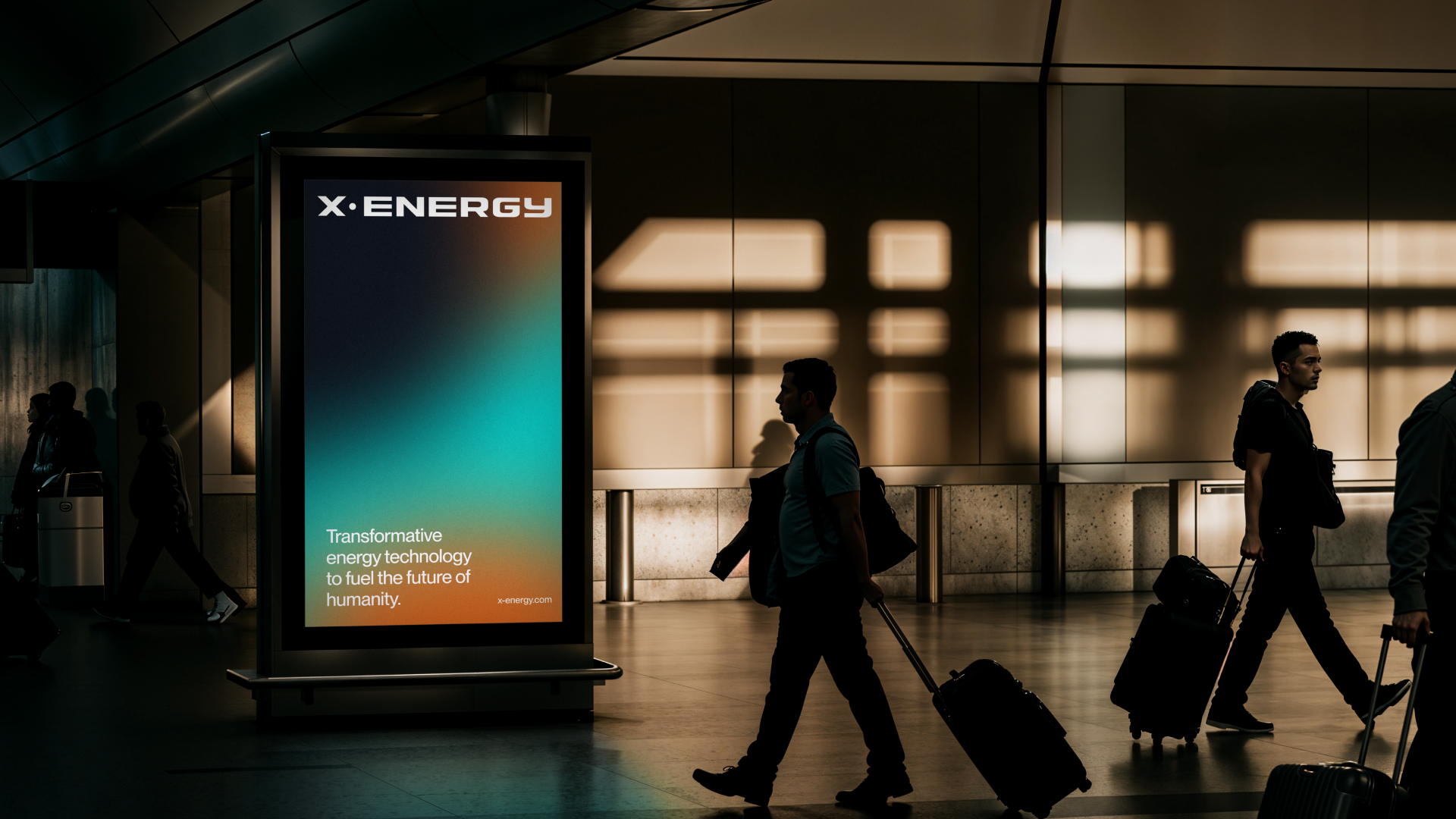 Silhouettes of travelers walking with rolling luggage past an illuminated advertisement for X:Energy at an airport. The ad features a gradient background and the words "Transformative energy technology to fuel the future of humanity," with the websit
