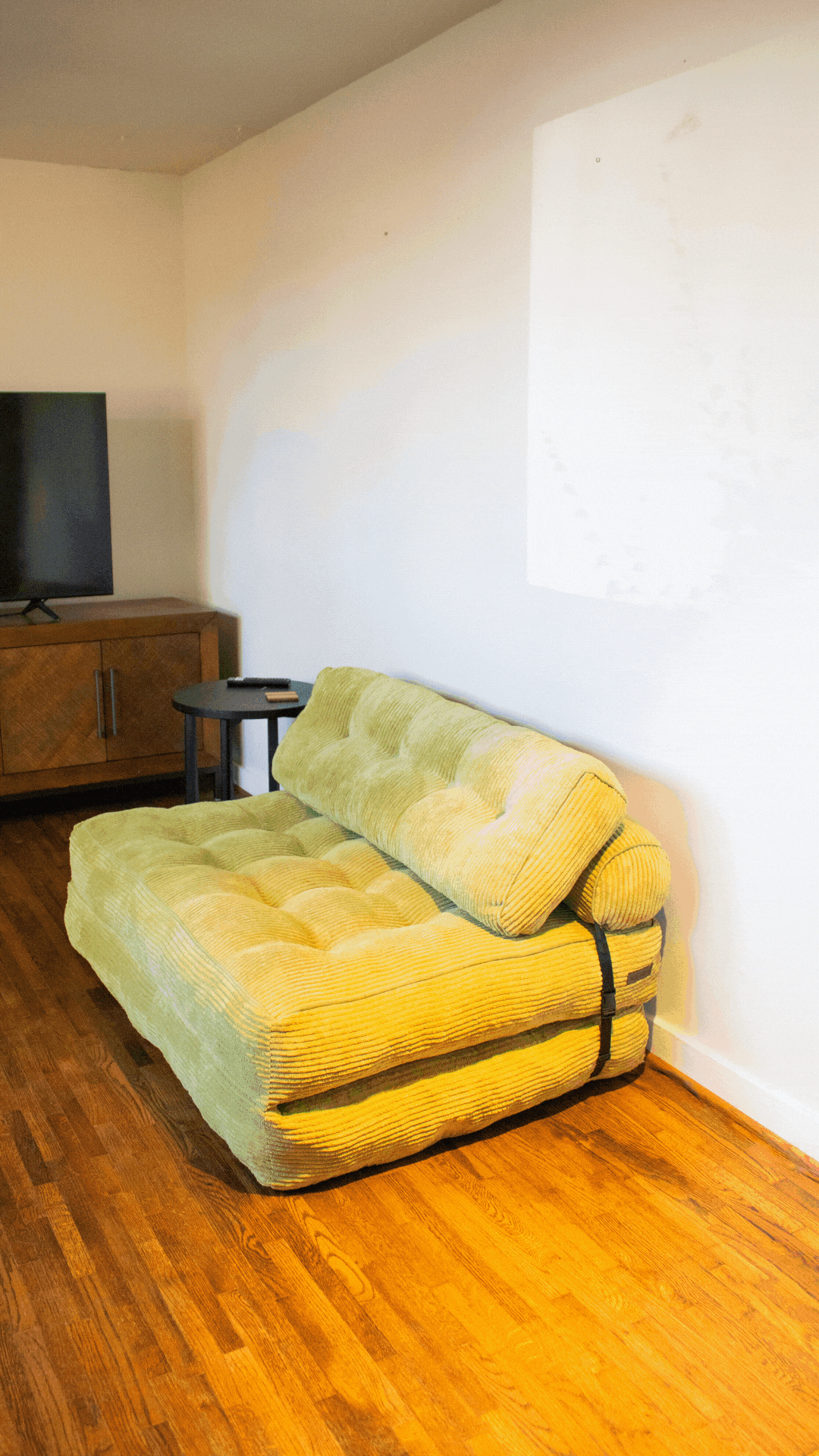 A green, corduroy sofa with a pillow backing, placed against a white wall in a living room. There is a wooden cabinet with a TV on top and a small black side table with a remote on it. The room has wooden flooring and a light-colored ceiling.