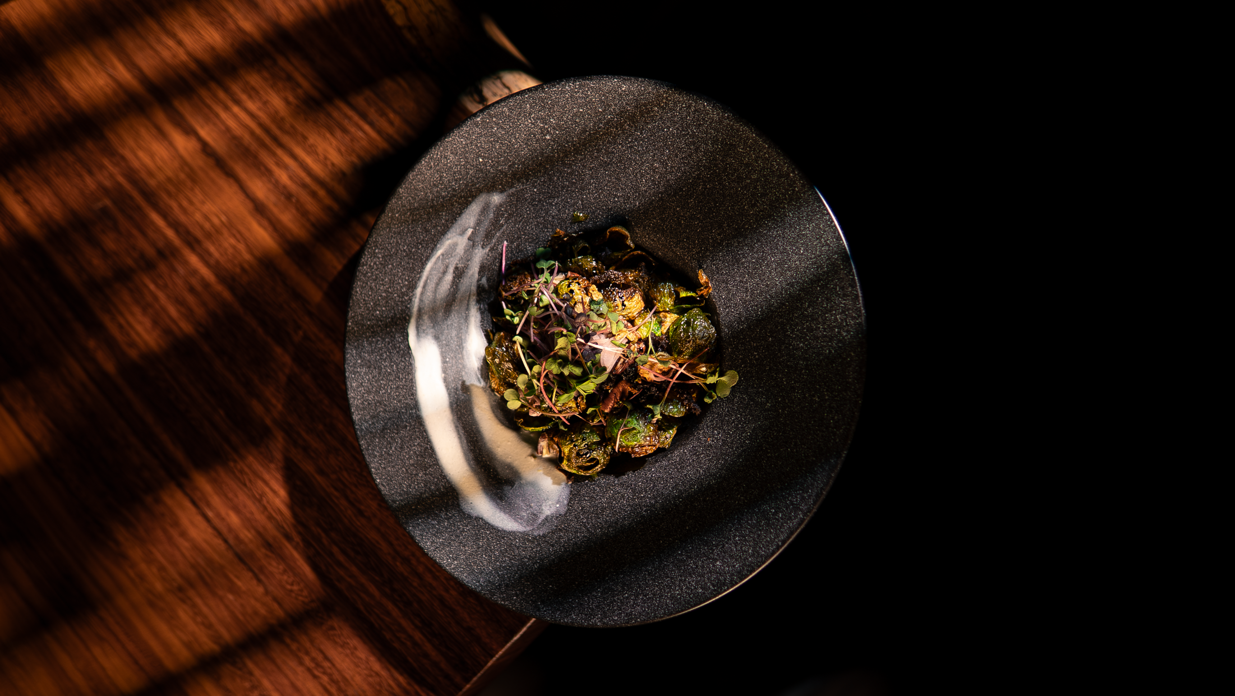 A plated gourmet dish with greens, microgreens, and a dark sauce on a dark, textured plate, set on a wooden table.