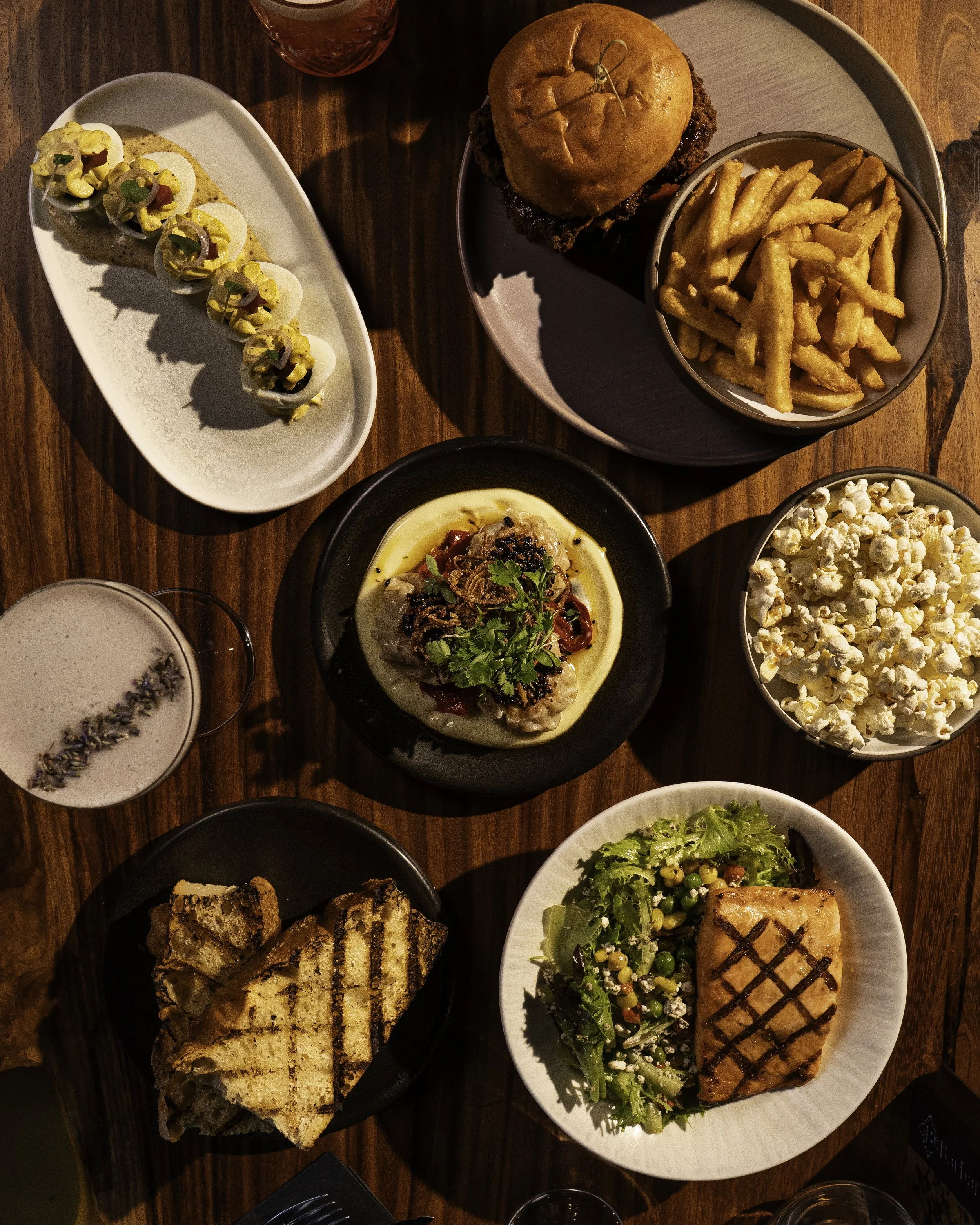 A table with various dishes including deviled eggs, a burger with fries, popcorn, a piece of grilled salmon with salad, a bread pudding, a bowl of chicken and dumplings, and a drink with lavender garnish.