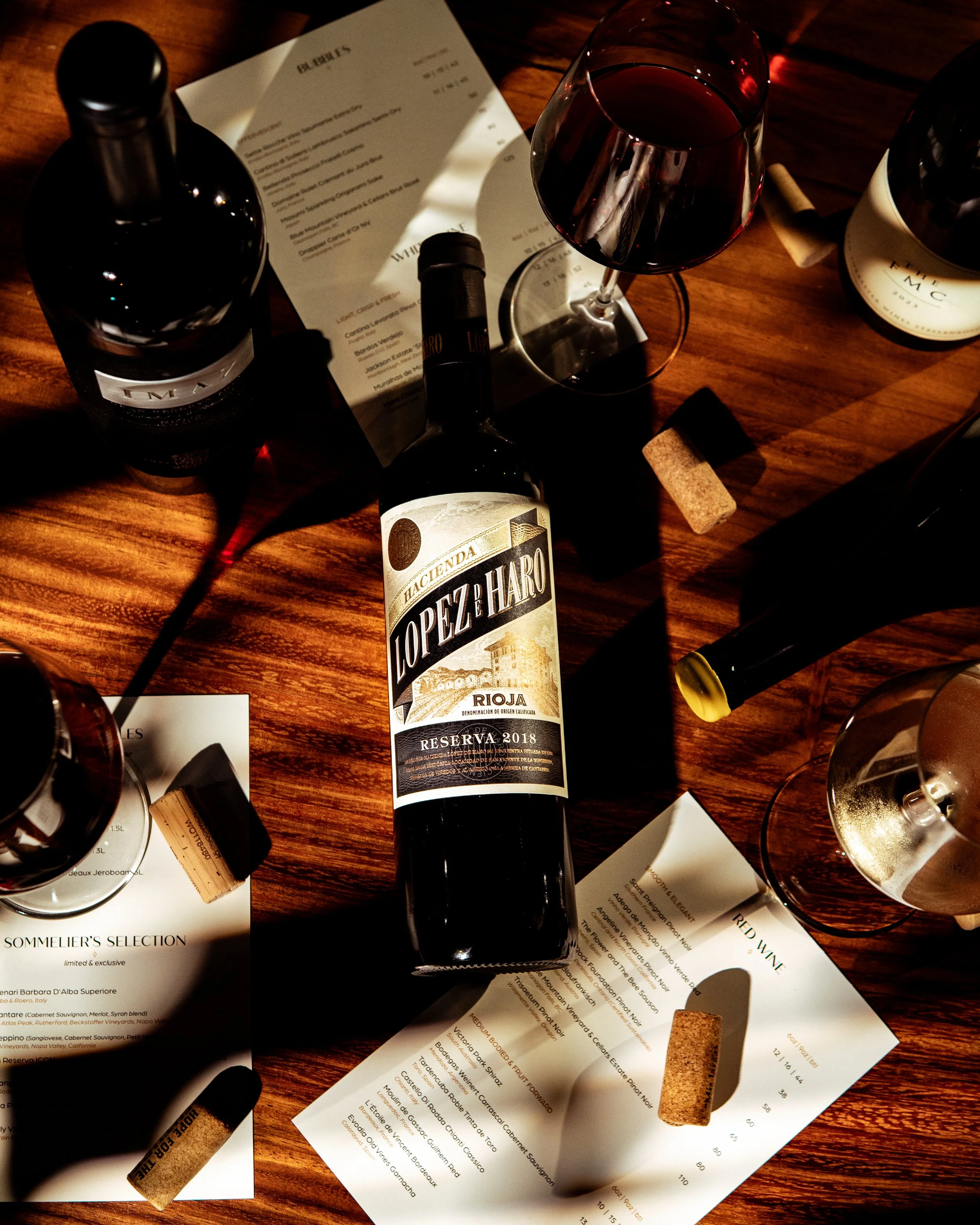 A bottle of Lopez de Haro Rioja Reserva 2018 wine, a filled wine glass, wine corks, a wine bottle opener, a wine bottle, and tasting notes papers on a wooden table with shadows.