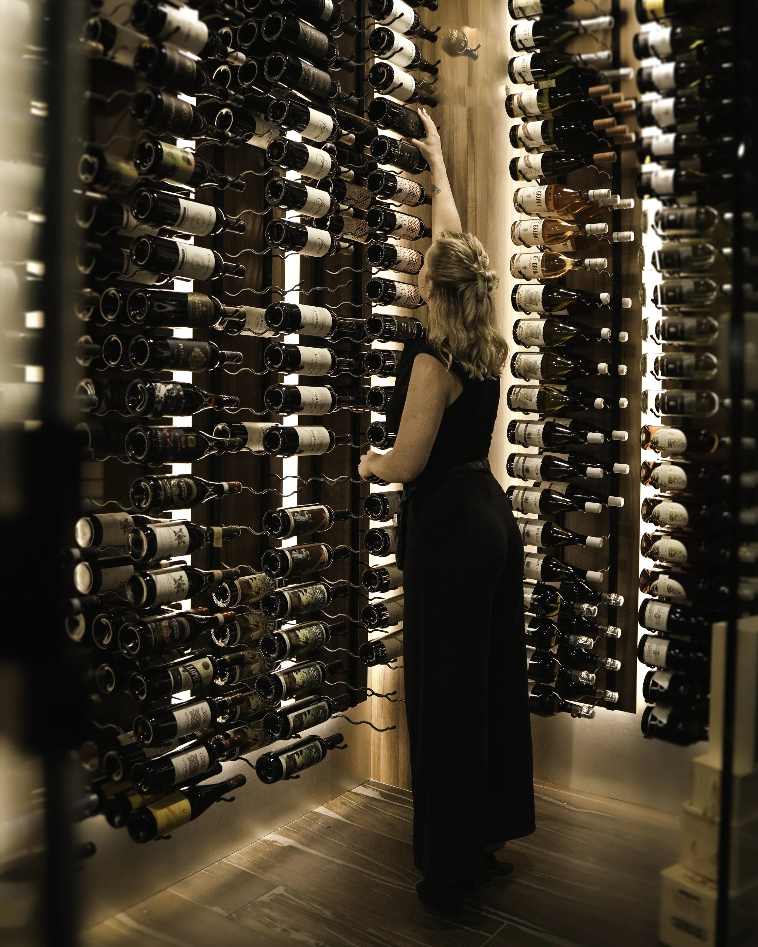 A woman looking at wine bottles stored on a wall in a wine cellar or store.