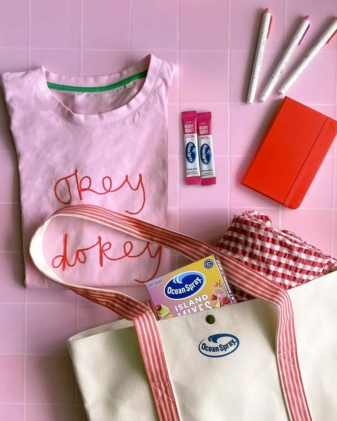 Ocean Spray Island Mixes box and two Berry Burst drink mix sticks beside a pink “okey dokey” embroidered t-shirt, an Ocean Spray tote bag, a red notebook and three pens on a pink tiled surface