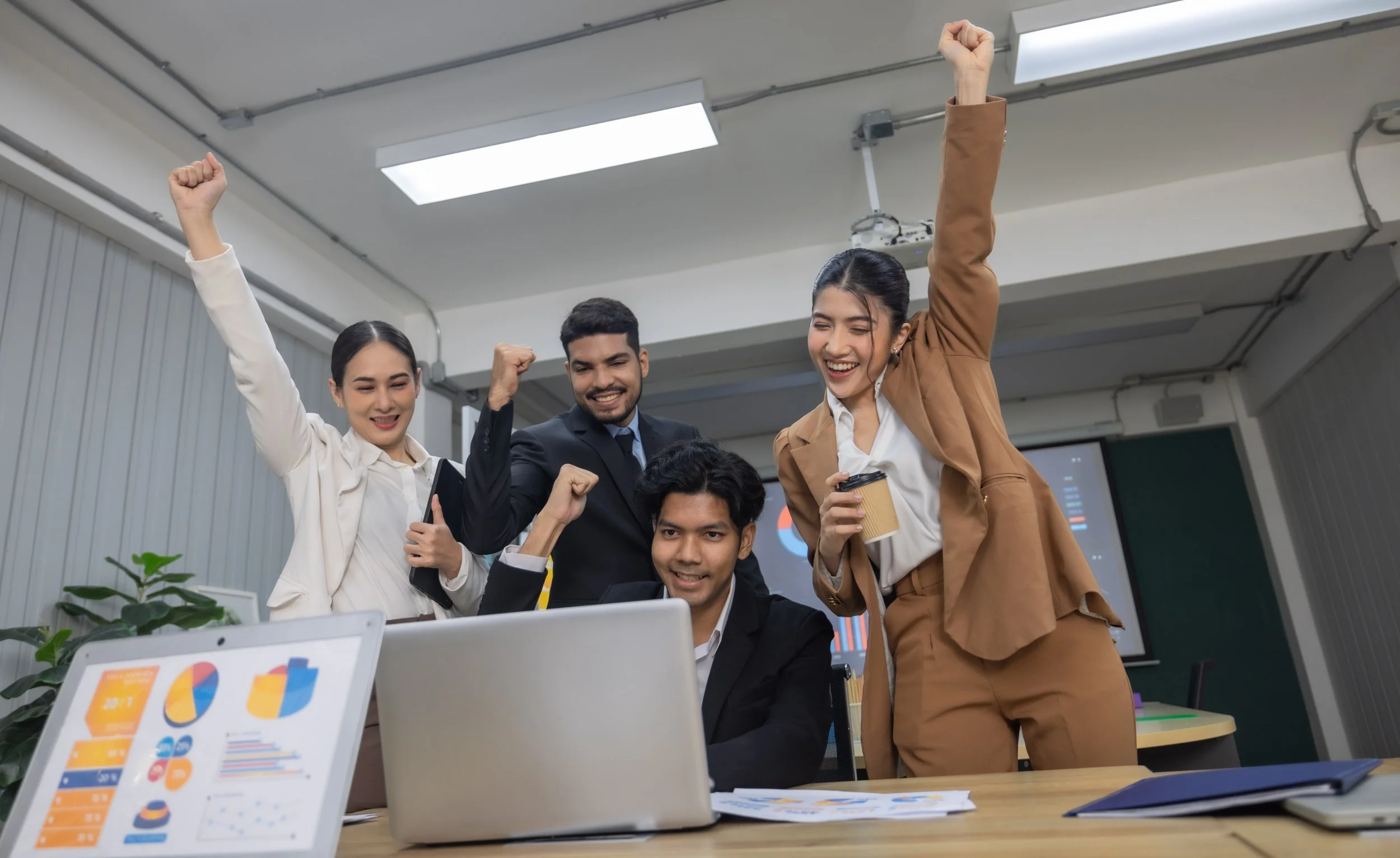 Four colleagues in an office celebrating with raised fists as they look at a laptop, with charts on a second screen and printed graphs on the desk.
