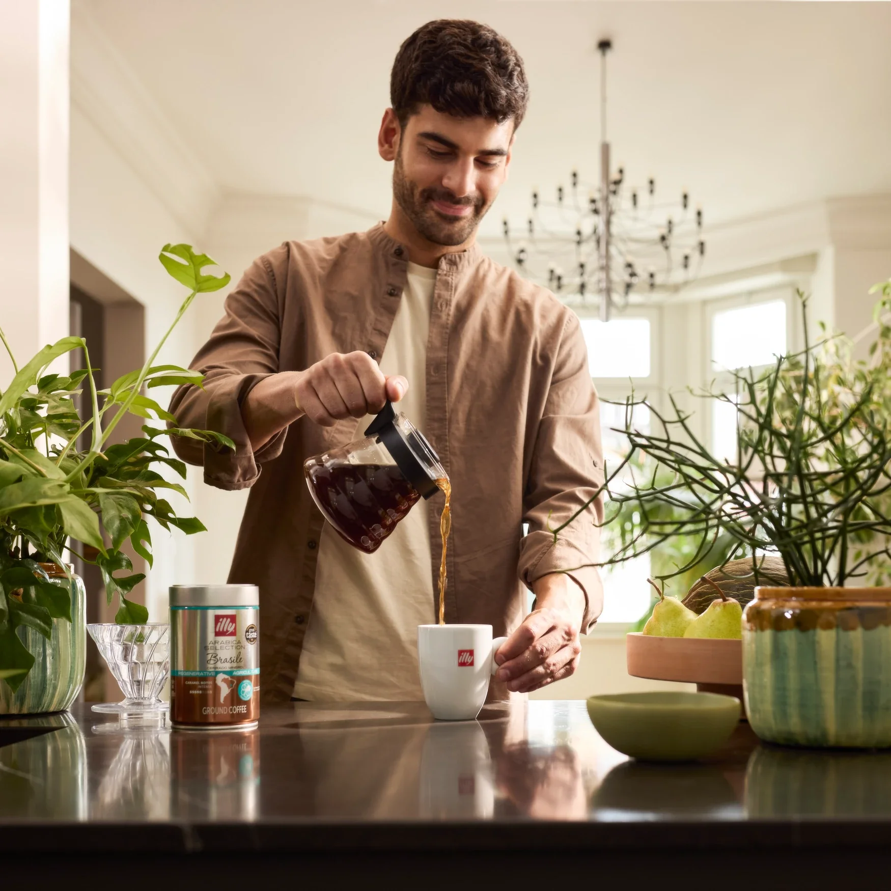 Smiling man in a brown shirt pouring filter coffee from a glass carafe into a white illy mug, beside a can of illy Arabica Selection Brasile ground coffee on a dark kitchen counter with green plants and pears