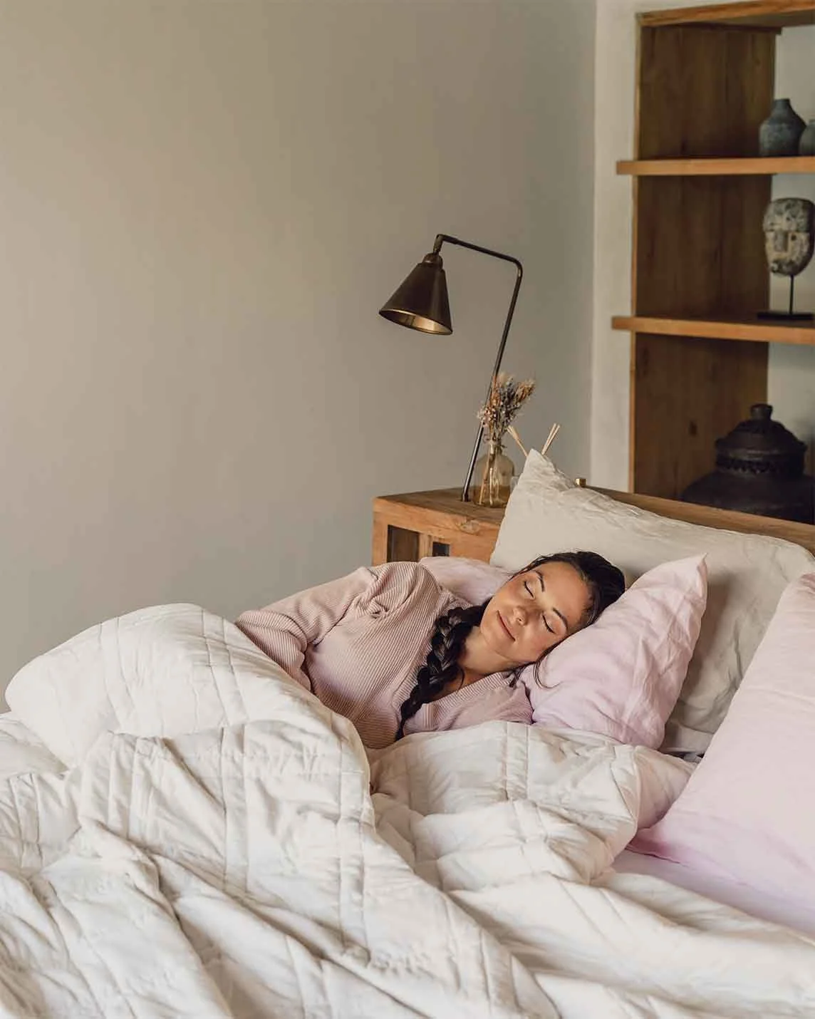 Woman with a long braid sleeping on a pink pillow under a cream quilted weighted blanket, beside a wooden bedside table with a brass lamp and wooden shelves