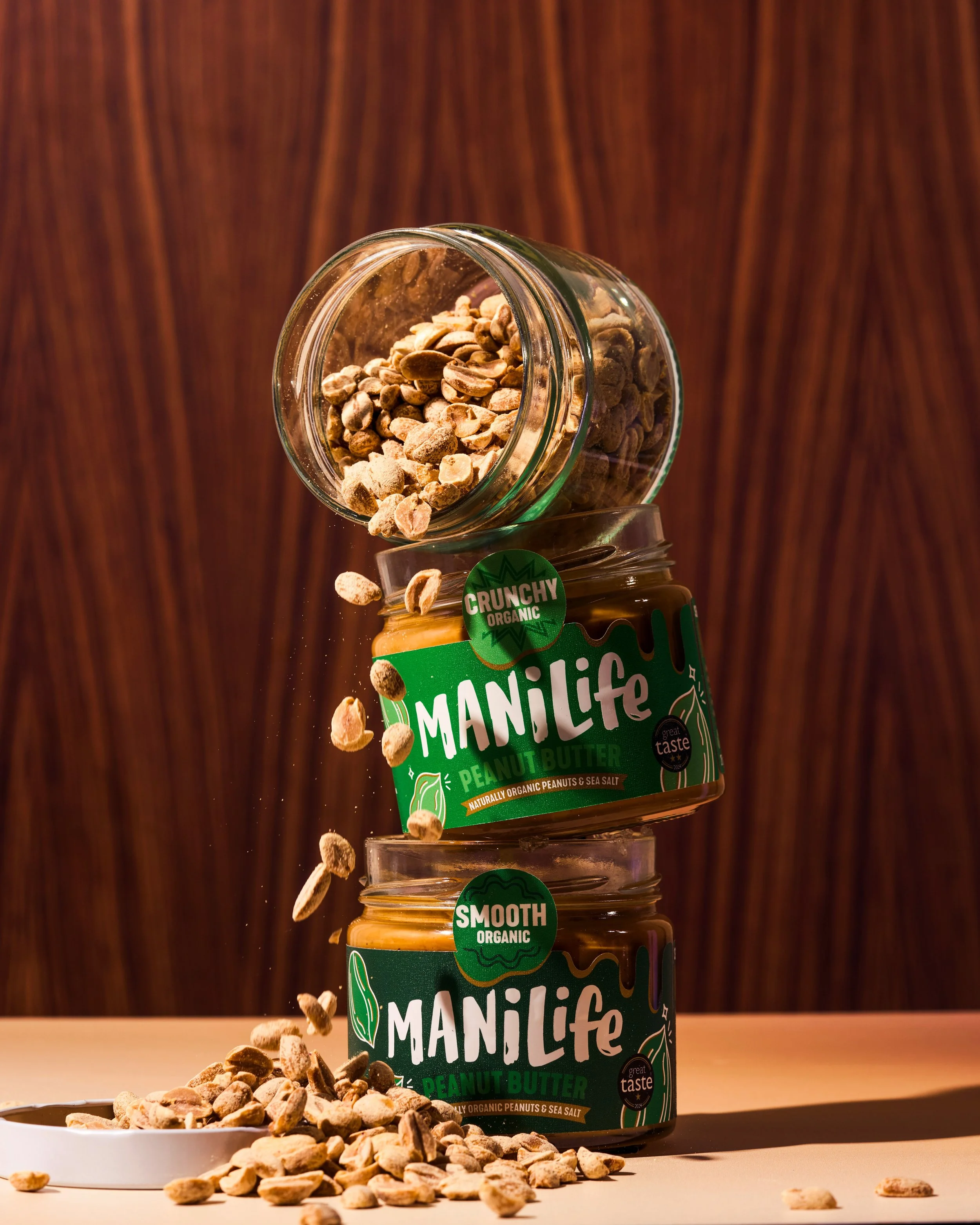 ManiLife Smooth Organic and Crunchy Organic peanut butter jars stacked on a wooden backdrop, with an open jar of salted peanuts tipping over them and peanuts falling onto the surface