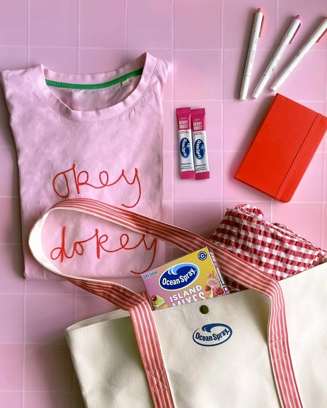 Ocean Spray Island Mixes box and two Berry Burst drink mix sticks beside a pink “okey dokey” embroidered t-shirt, an Ocean Spray tote bag, a red notebook and three pens on a pink tiled surface