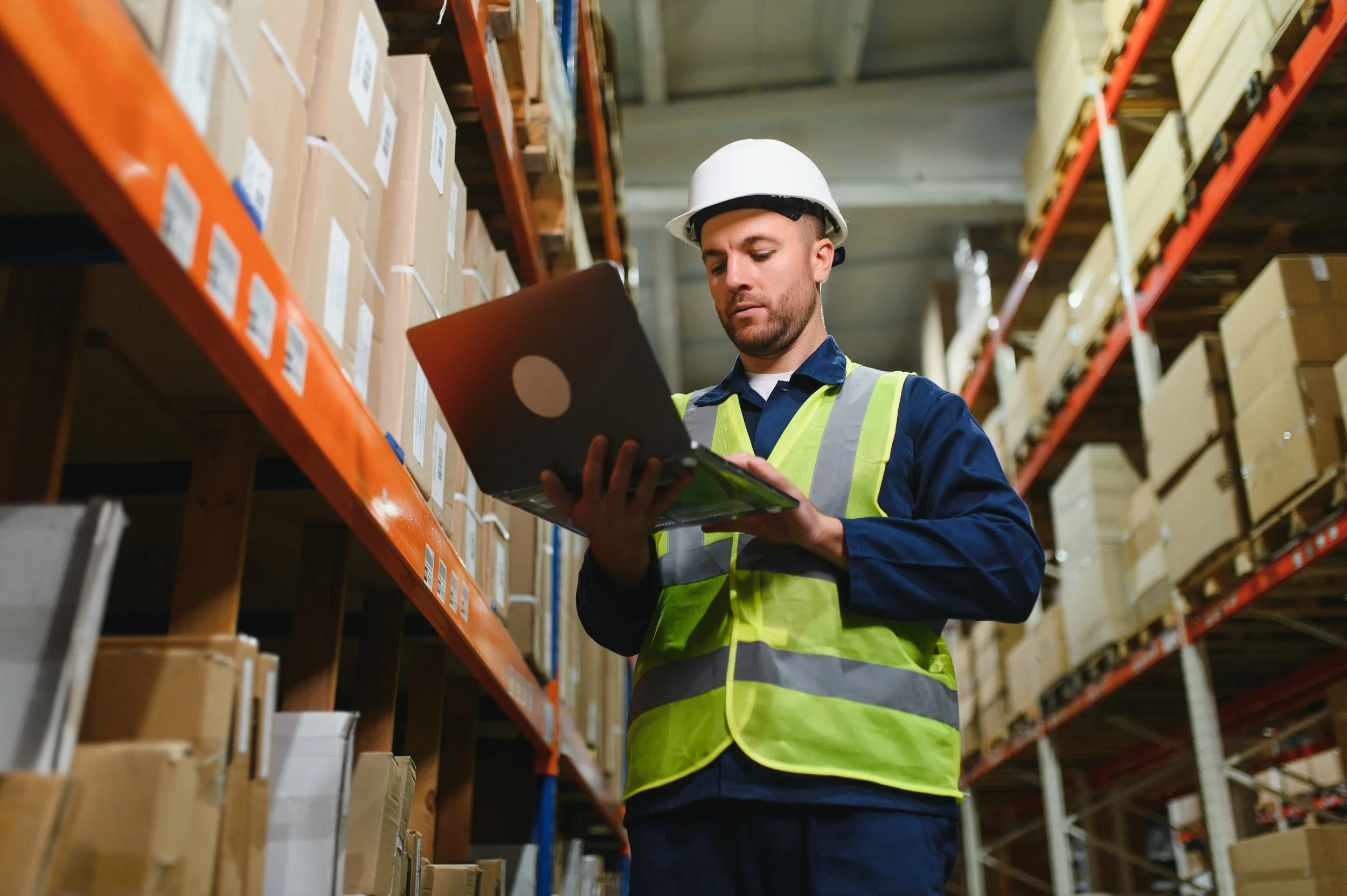 Warehouse worker in a hard hat and hi-vis vest checking inventory on a laptop between rows of shelved boxes.