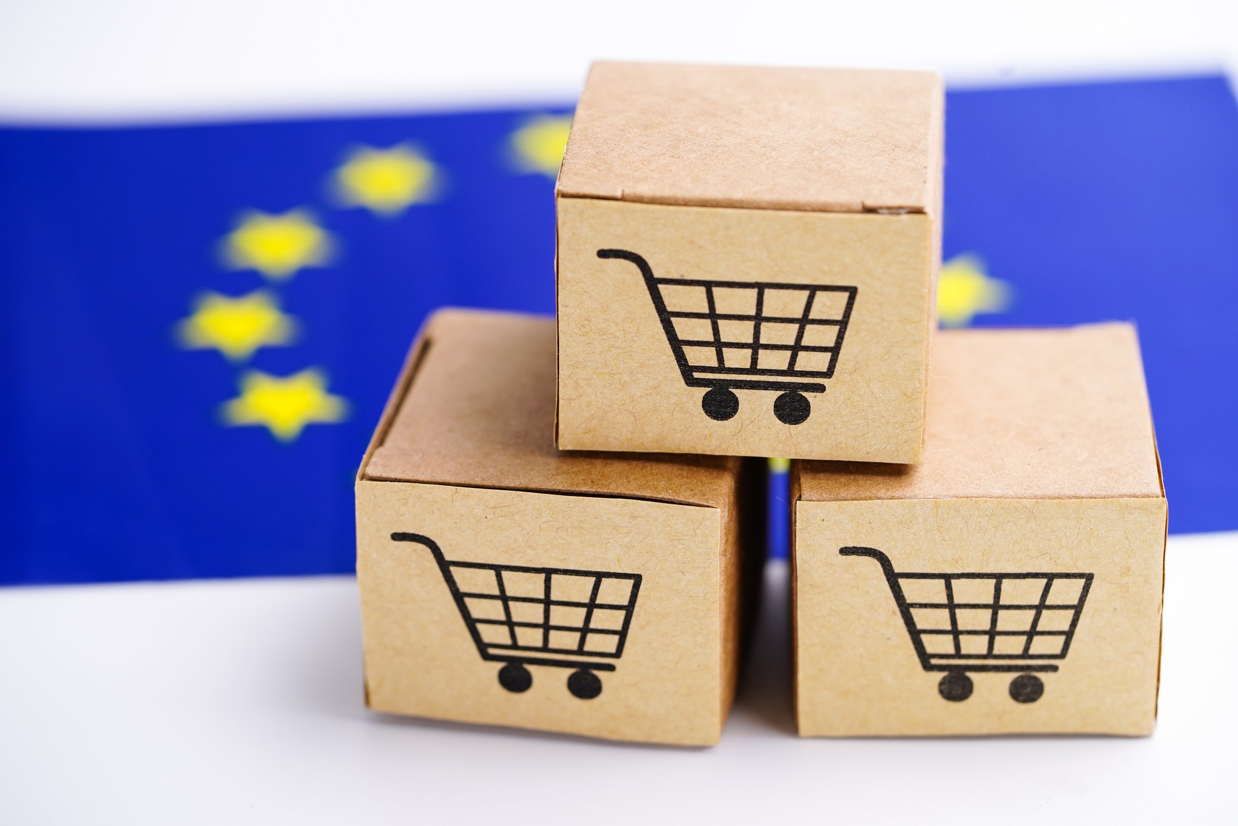 Three small cardboard boxes with shopping cart icons stacked in front of a European Union flag