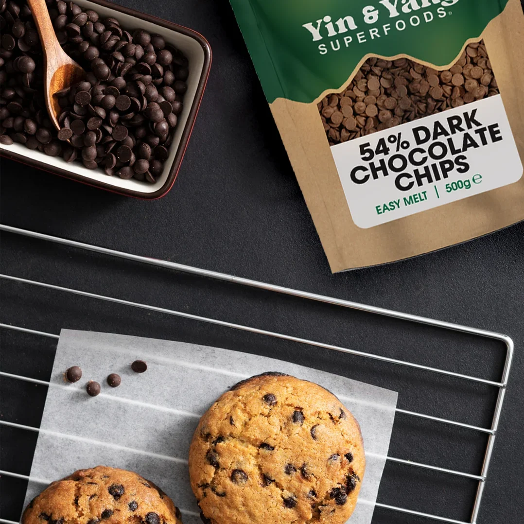 Yin & Yang Superfoods 54% dark chocolate chips bag beside a bowl of chocolate chips and a chocolate chip cookie on a cooling rack