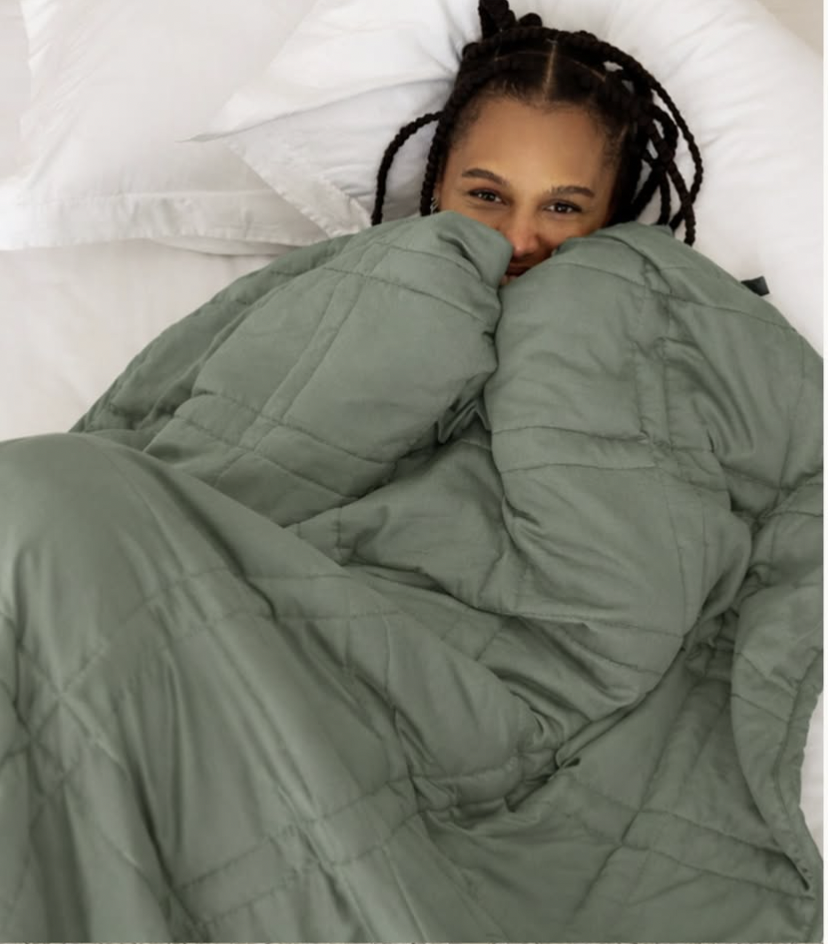 Smiling woman with braids lying in bed on white pillows, pulling a sage green quilted weighted blanket up to her nose