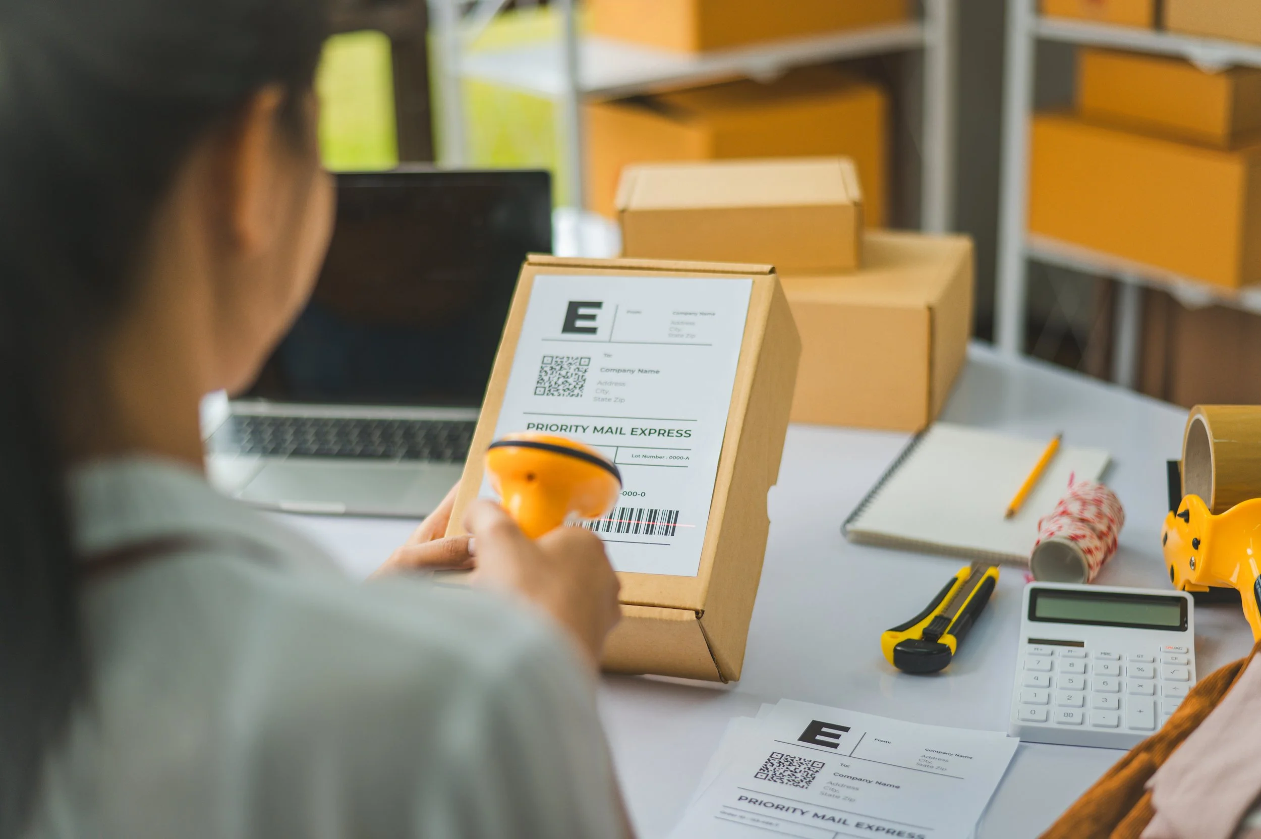 Person scanning the barcode on a shipping label stuck to a cardboard parcel at a packing desk, with stacked boxes, a laptop, calculator, notebook and tape nearby.