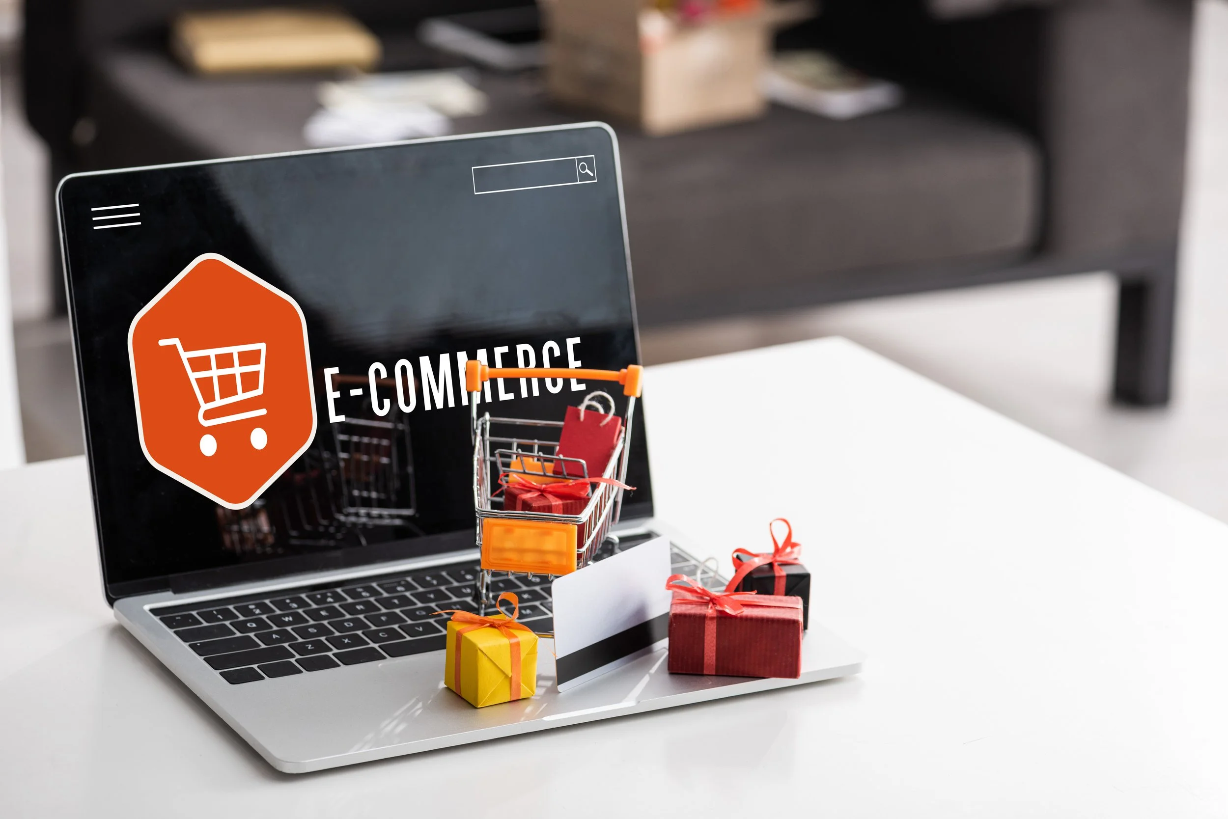 Laptop displaying an "E-commerce" screen with a shopping cart icon, surrounded by a small shopping cart, wrapped gift boxes, and a credit card on the keyboard