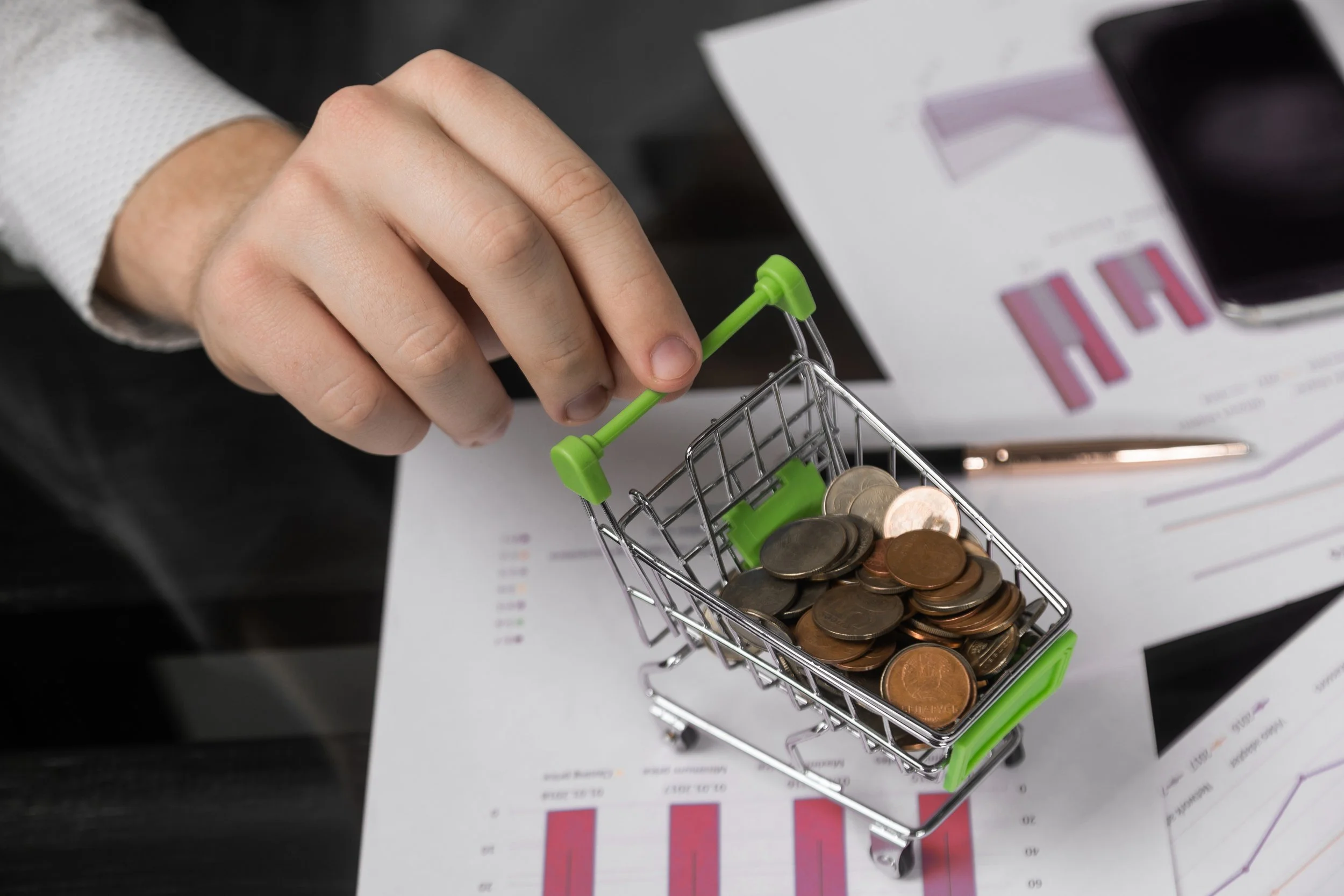 Businessman holding a small shopping cart filled with coins over printed financial charts and graphs, with a smartphone and pen in the background