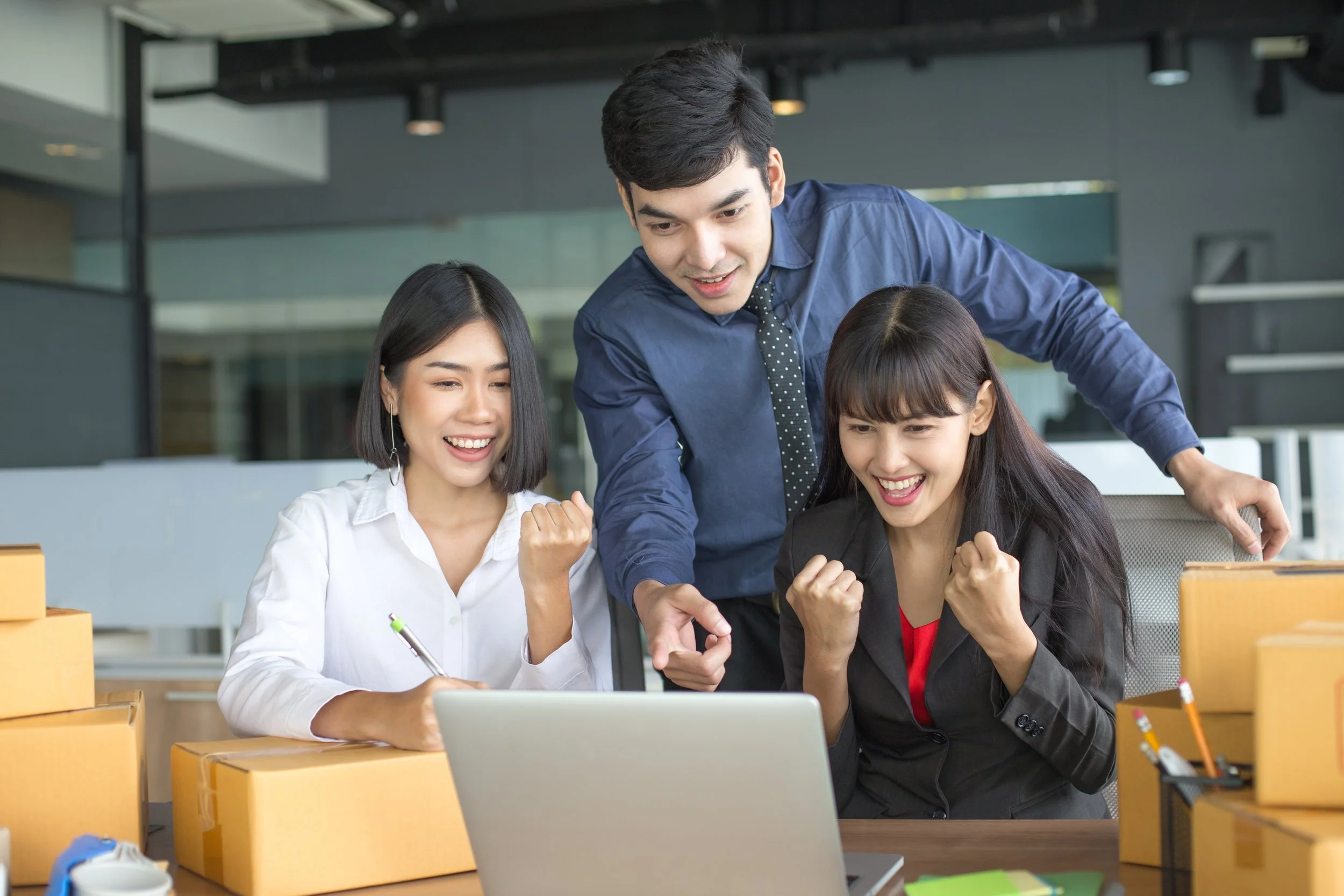 Three coworkers celebrating with clenched fists while looking at a laptop screen, surrounded by cardboard shipping boxes in an office