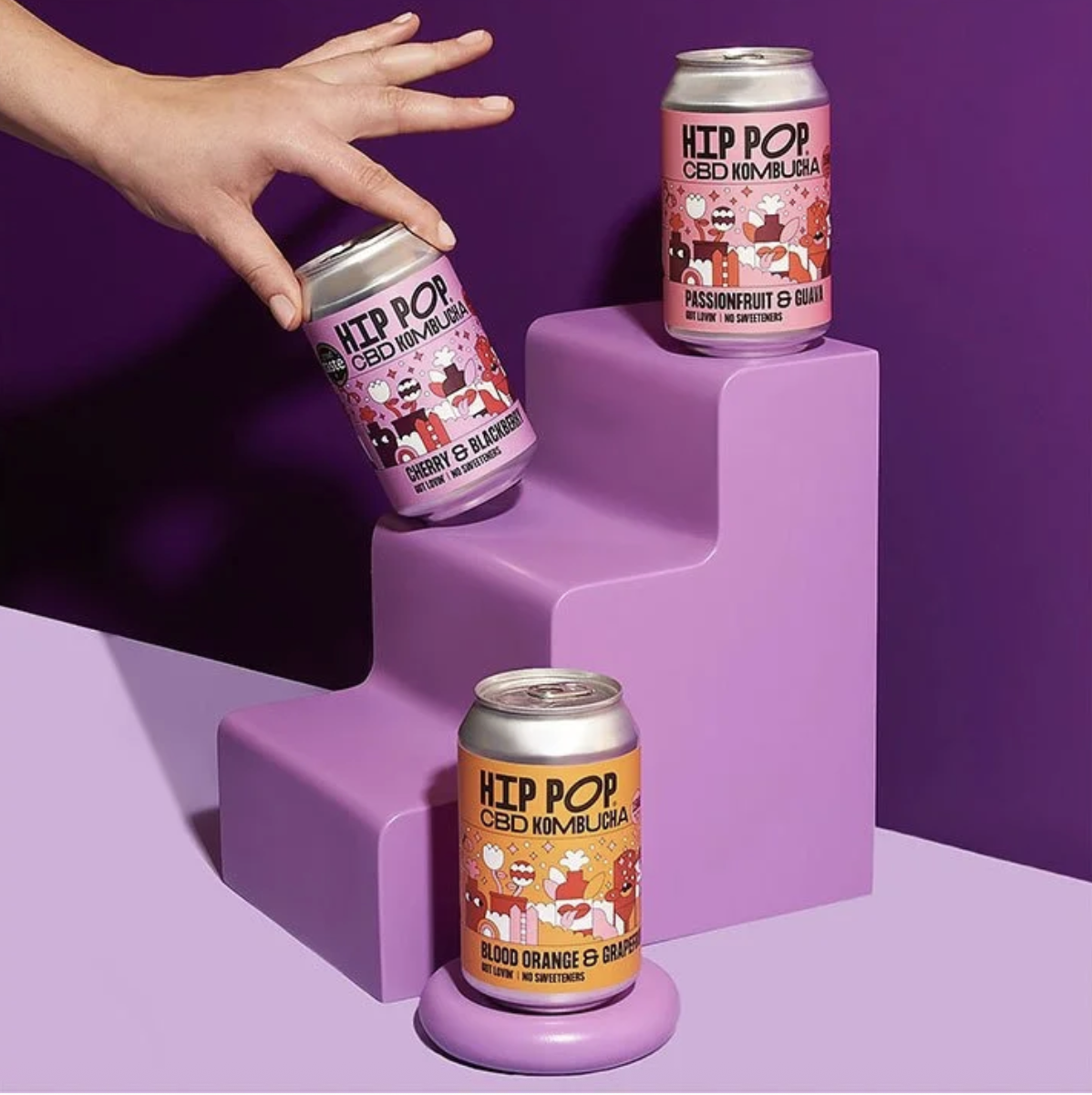 Hip Pop CBD Kombucha cans in Cherry & Blackberry, Passionfruit & Guava and Blood Orange & Grapefruit flavours, styled on purple podiums with a hand reaching for one can
