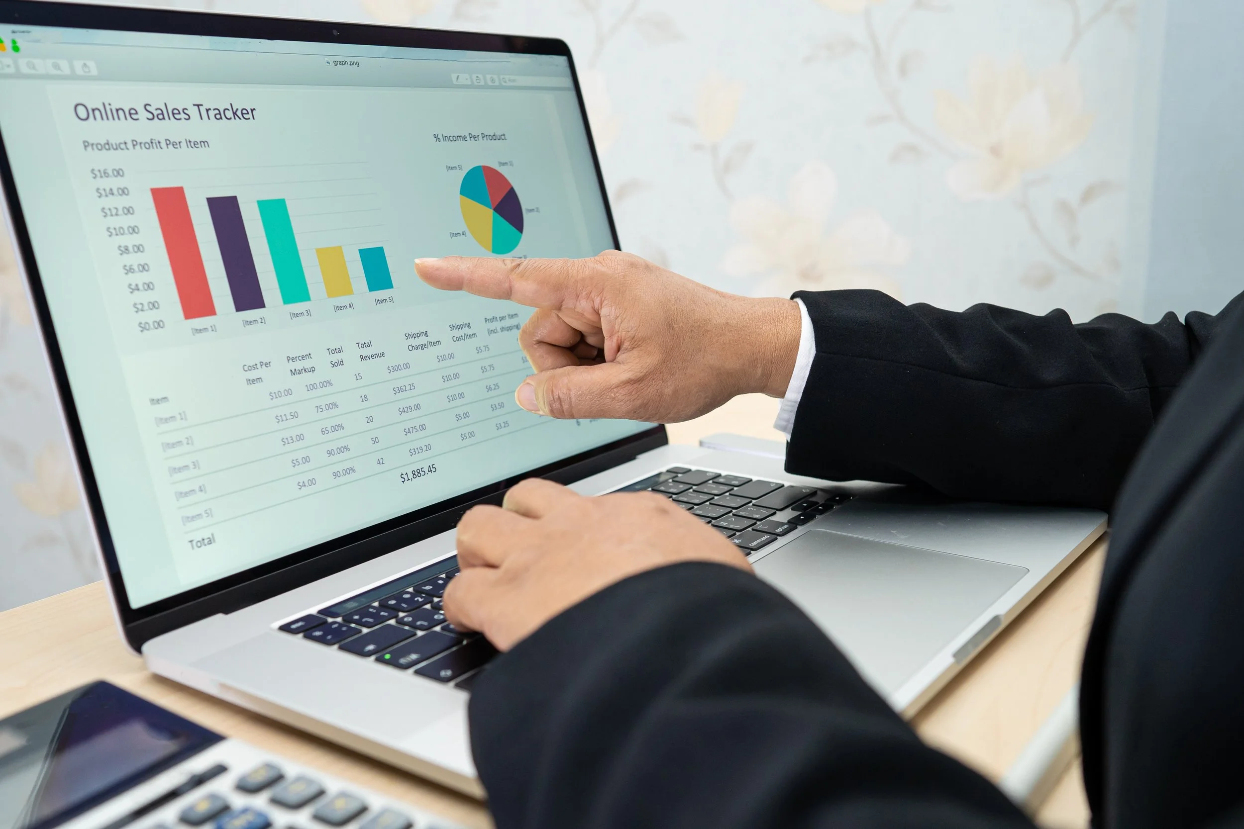 Hand pointing at a laptop screen showing an online sales tracker with a bar chart, pie chart, and product profit table