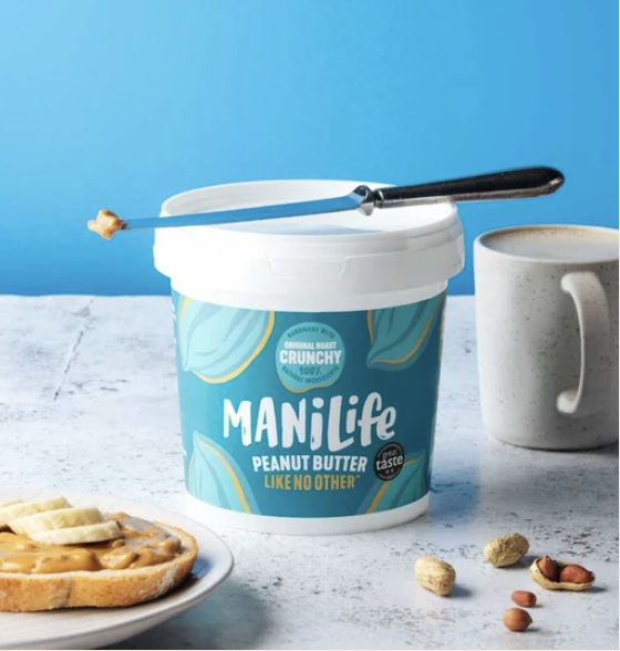 ManiLife Crunchy peanut butter tub with a knife resting across the lid, beside a slice of toast with peanut butter and banana, a mug of coffee and a scatter of whole peanuts