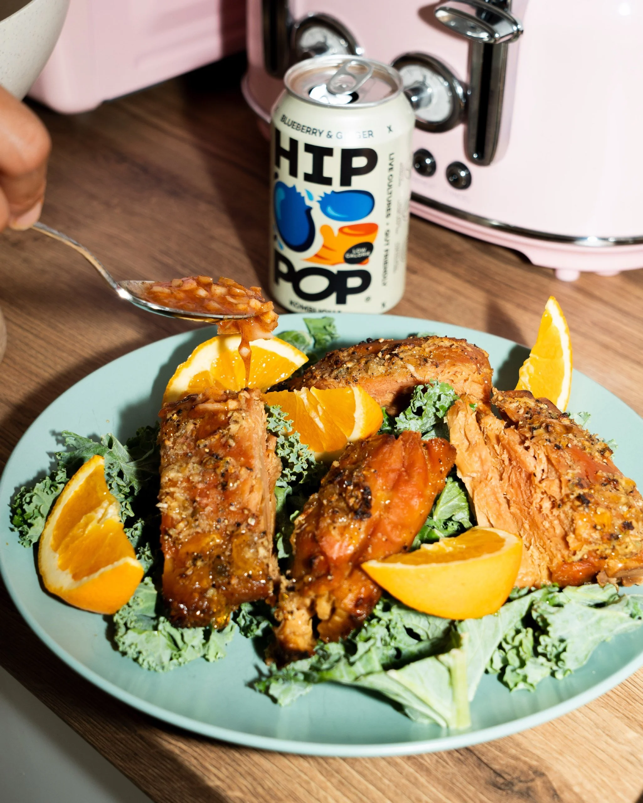 Glazed salmon pieces with orange wedges on a bed of kale on a blue plate, a spoon of sauce held above it and a can of Hip Pop Blueberry & Ginger kombucha beside a pink toaster