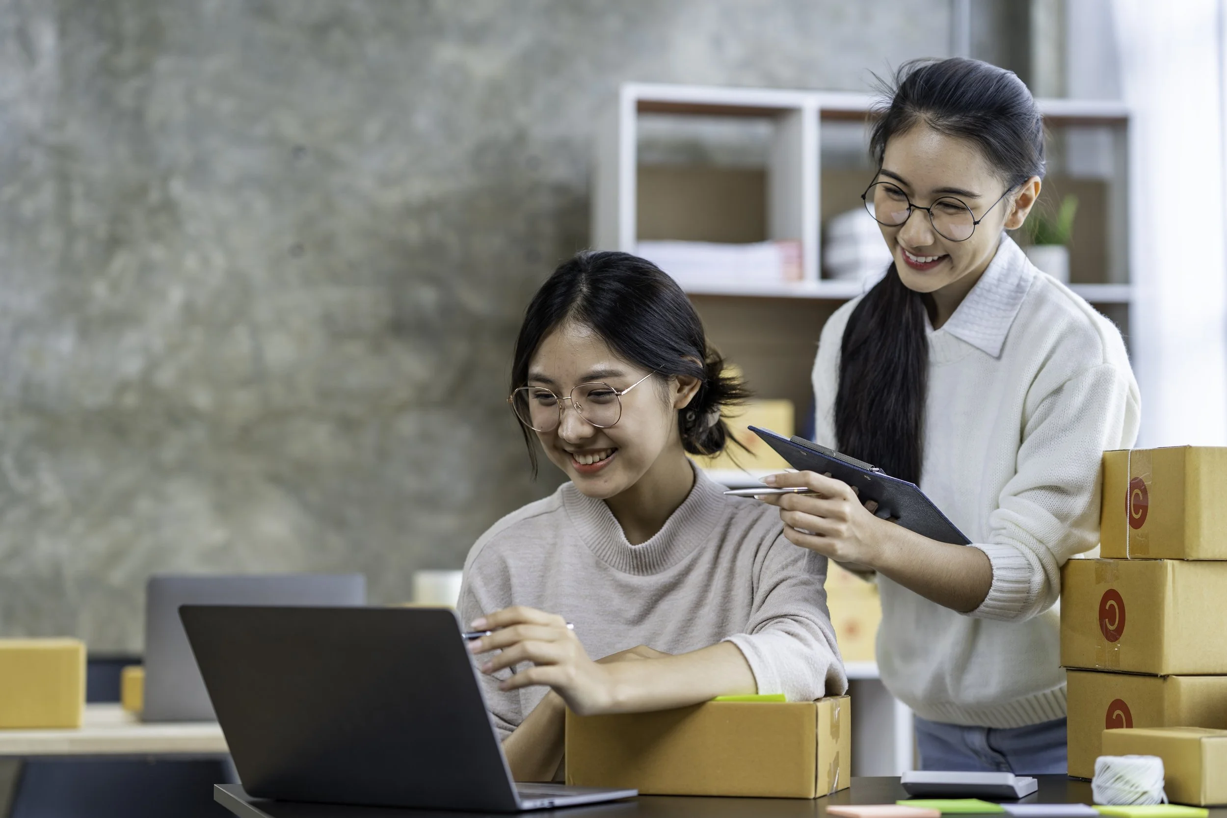 Two sellers reviewing a new product launch on a laptop, surrounded by inventory boxes ready for FBA
