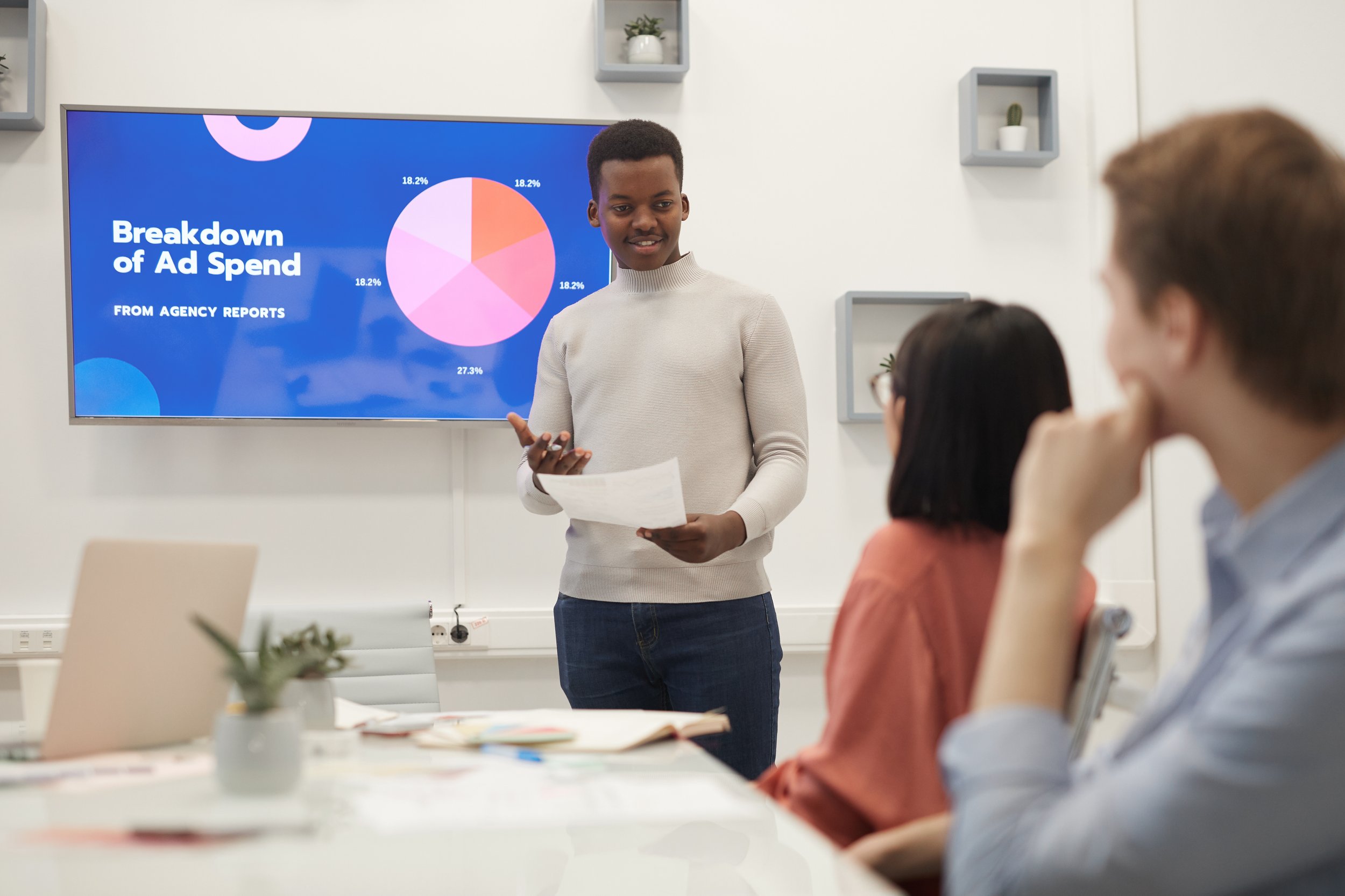 Presenter showing a team an ad spend breakdown pie chart on a screen during a meeting.