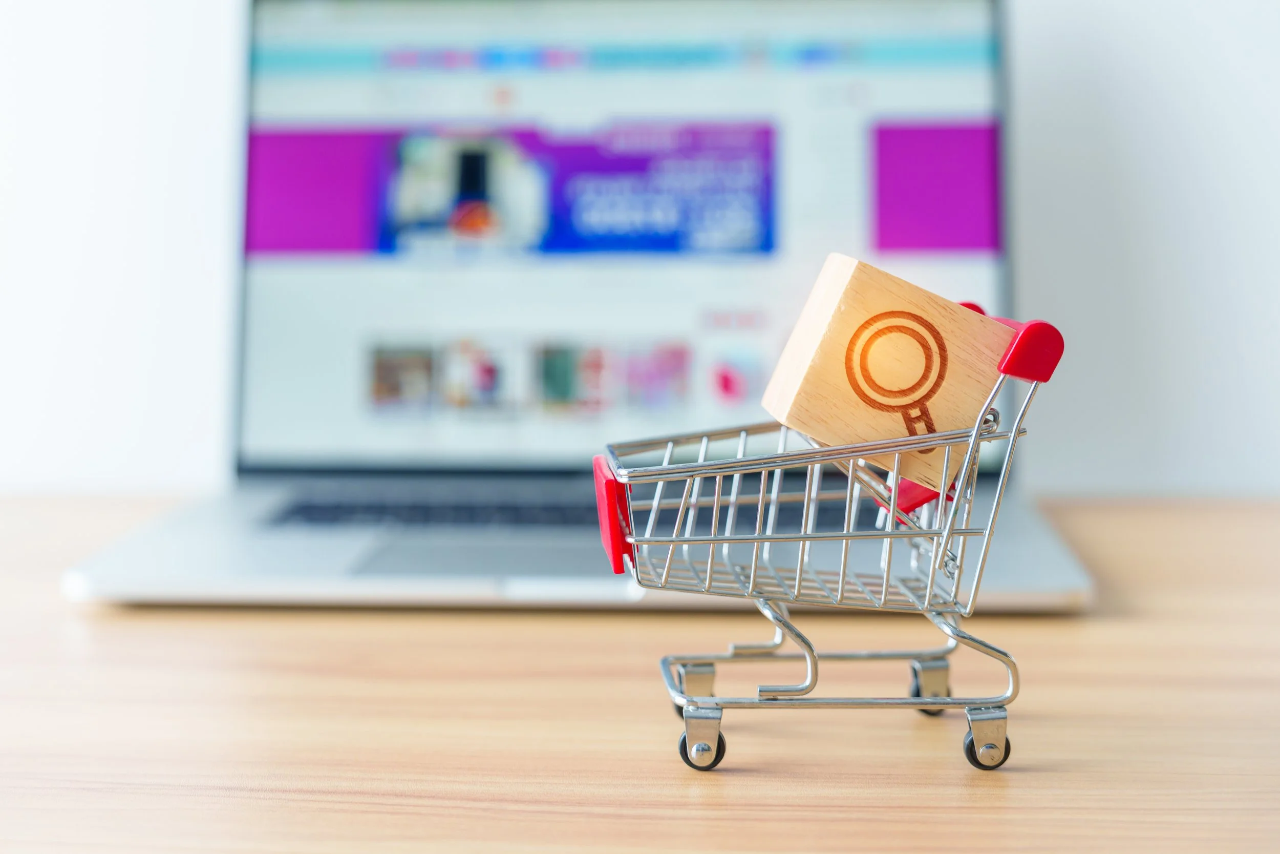 Wooden cube with a search icon inside a small shopping cart in front of a laptop showing an online store