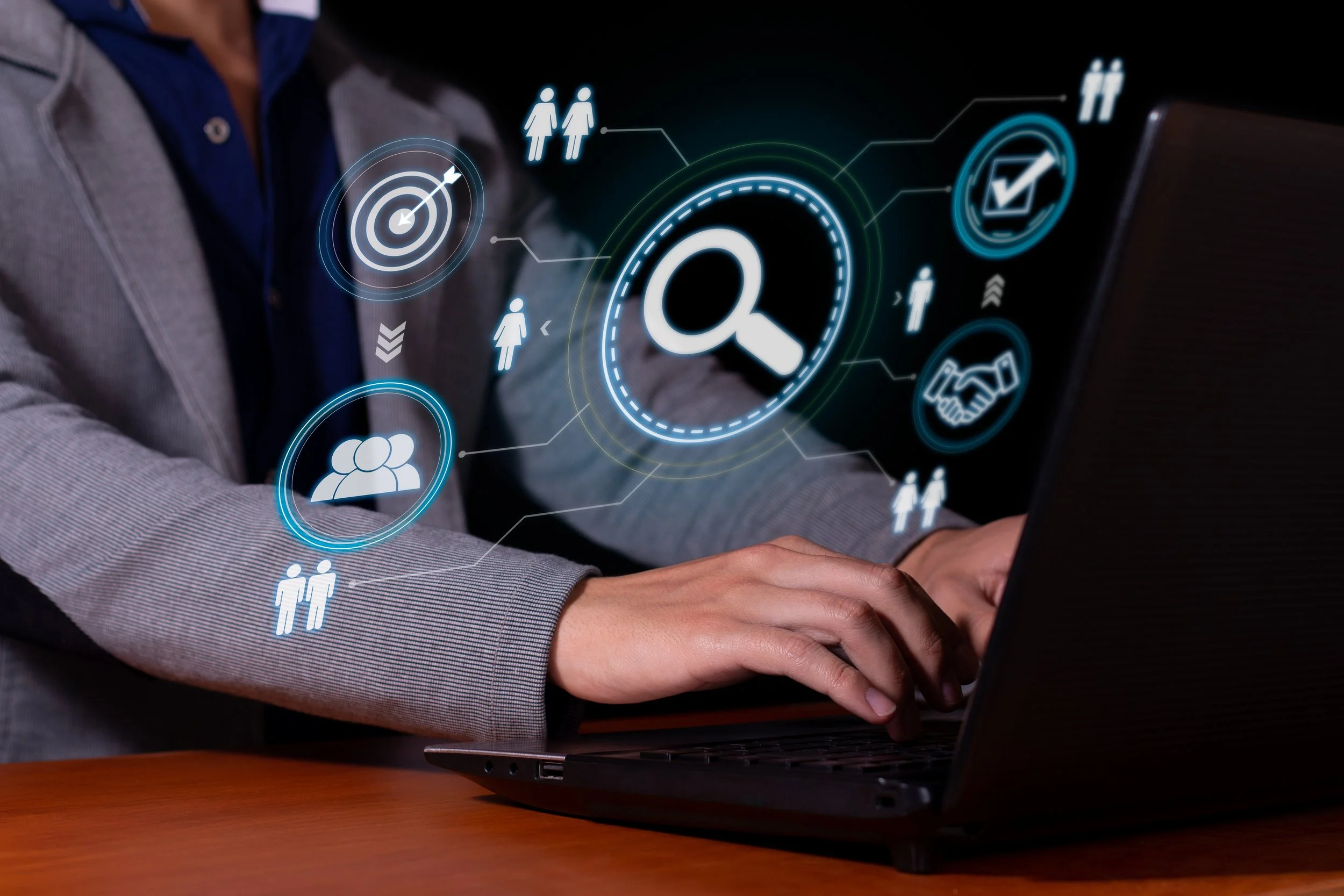 Person in a grey blazer typing on a laptop with glowing icons floating above, including a magnifying glass, target, group of people, handshake and checkmark.