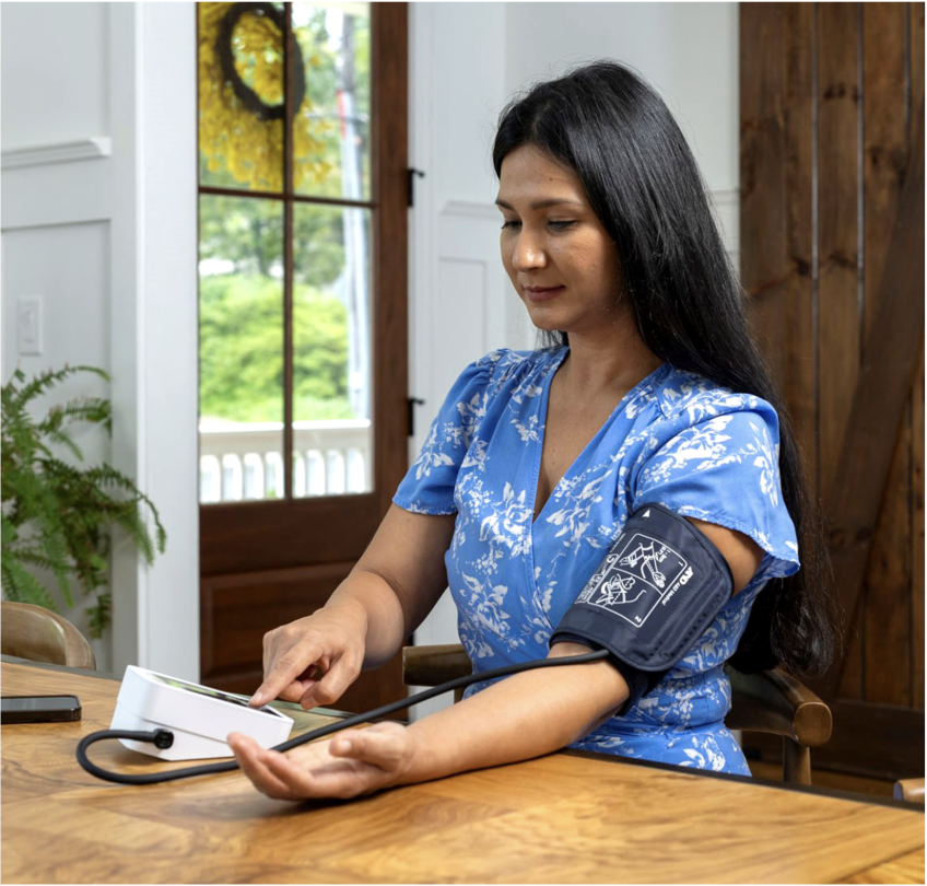 A woman seated at a wooden table checking her blood pressure at home, with an A&D Medical upper arm cuff on her left arm and her finger on the white monitor