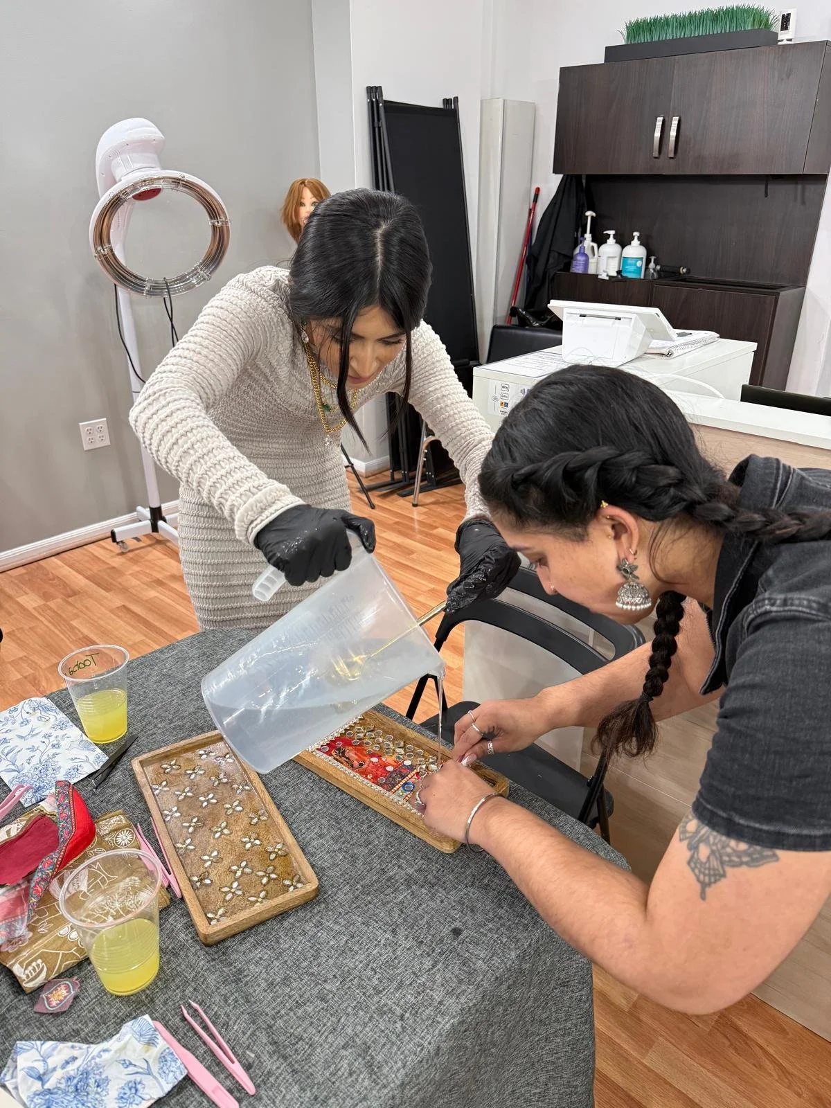 Two women working on jewelry projects at a table, with one pouring liquid and the other positioning jewelry components. The background includes a hair steamer, a mannequin head, and shelves with bottles.