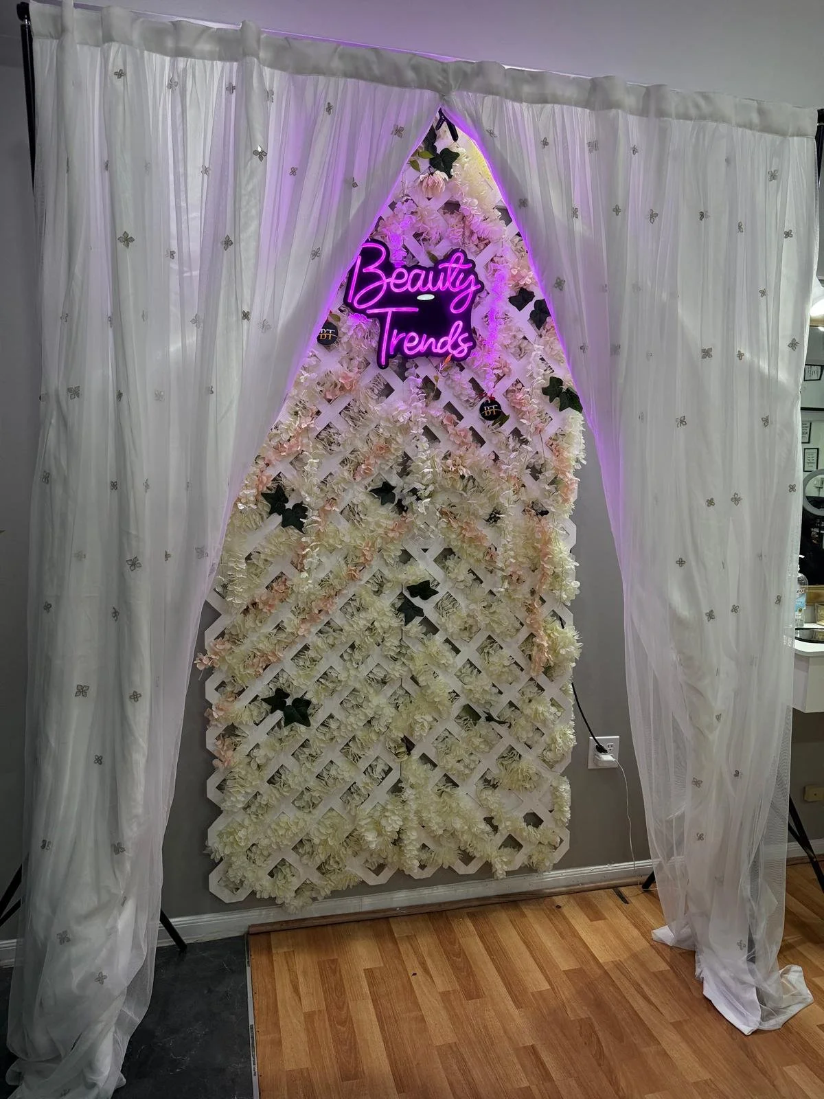 A decorative floral backdrop with a white lattice frame filled with white and light pink flowers, black leaves, and small white blooms. A pink neon sign reads 'Beauty Trends' and is mounted near the top. White curtains with small silver butterfly pat