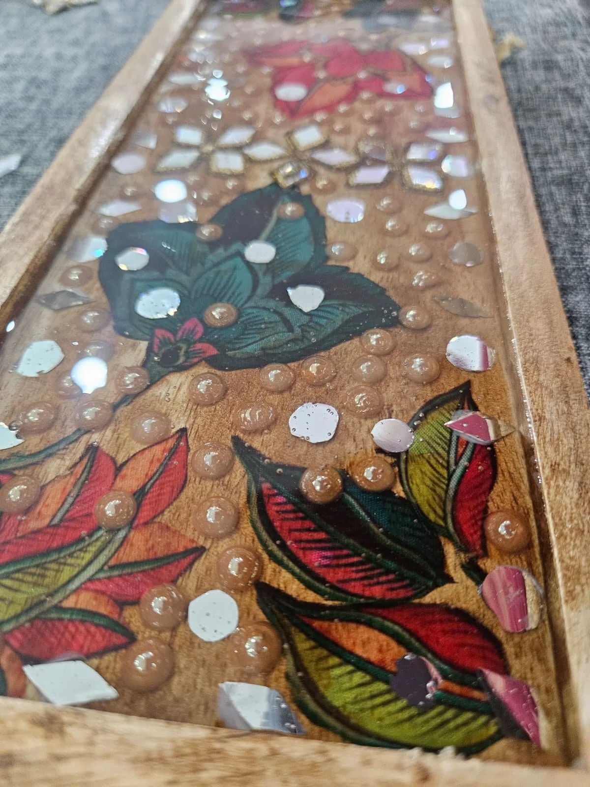 Close-up of a wooden artwork with floral designs and embedded sequins or beads under a glass cover.