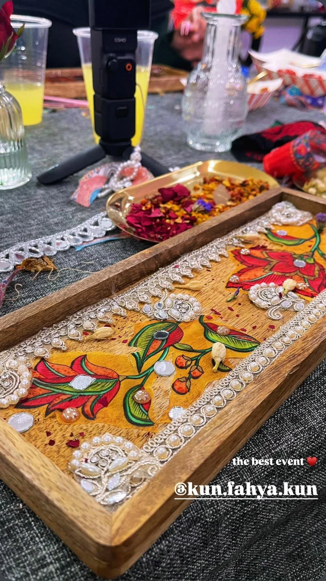 Decorative wooden tray with hanging ornaments, sequins, and colorful beads on a table with drinks and flowers.