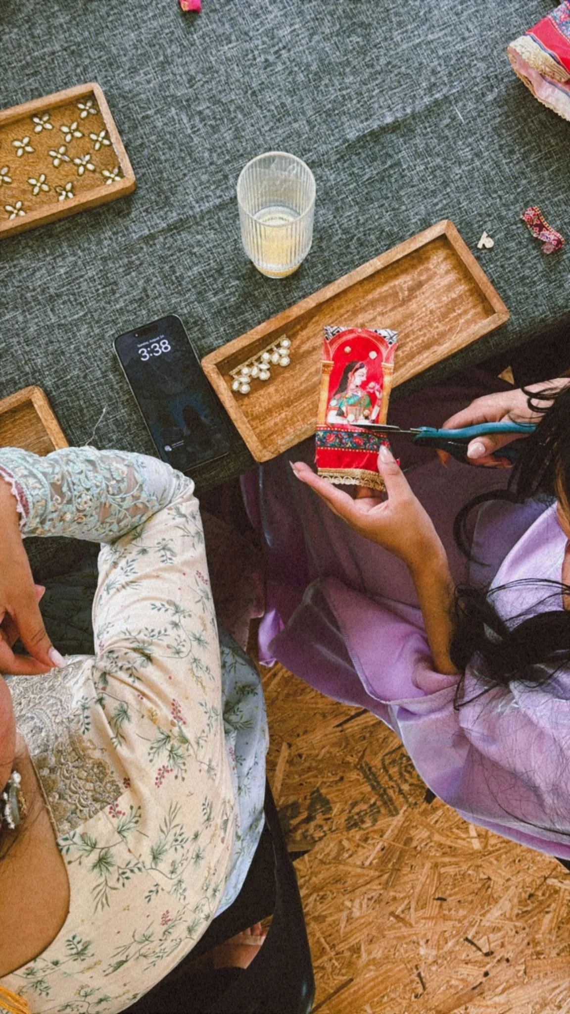 A person is using scissors to cut a decorative item with an image of a woman on it. The scene is at a table with wooden trays, a glass of water, and scattered small white seeds or beads. Another person, dressed in patterned clothing, is sitting nearb