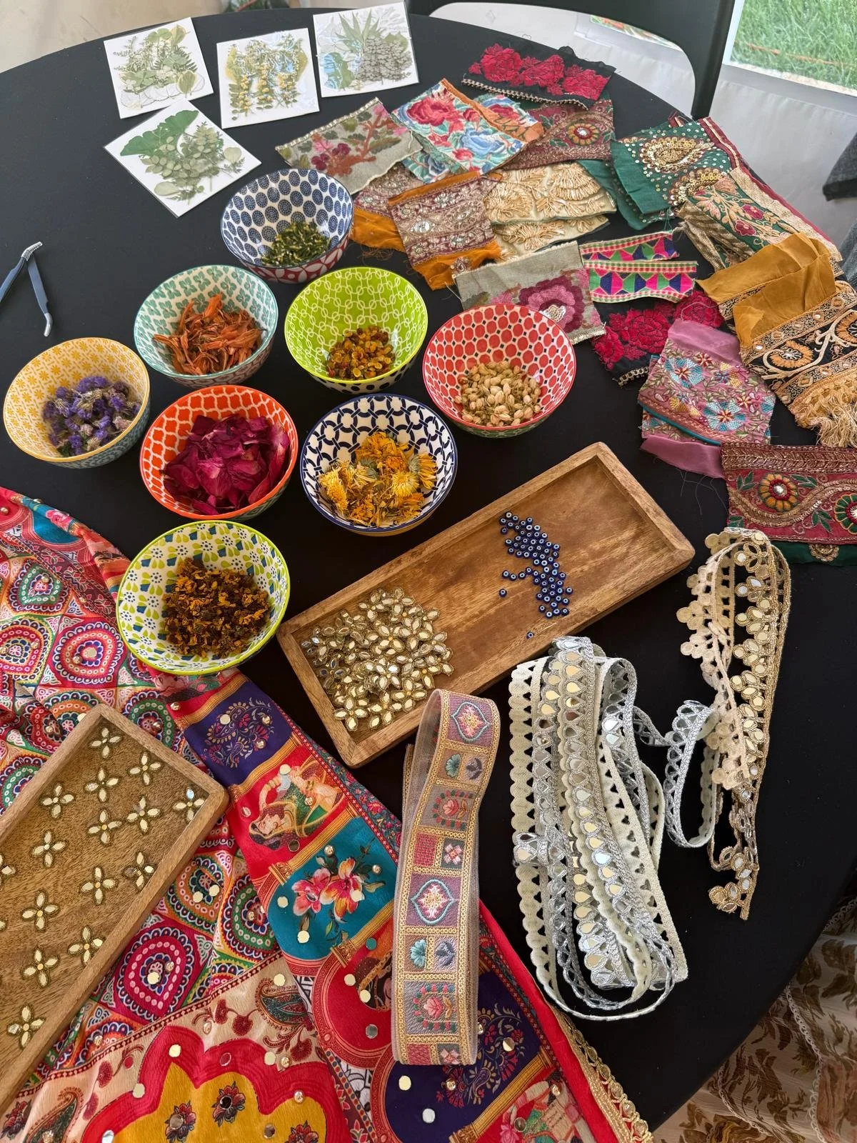Array of colorful embroidered fabrics, ribbons, and beads on a black table surrounded by bowls filled with dried flowers, herbs, and sequins, indicating a craft or textile project.