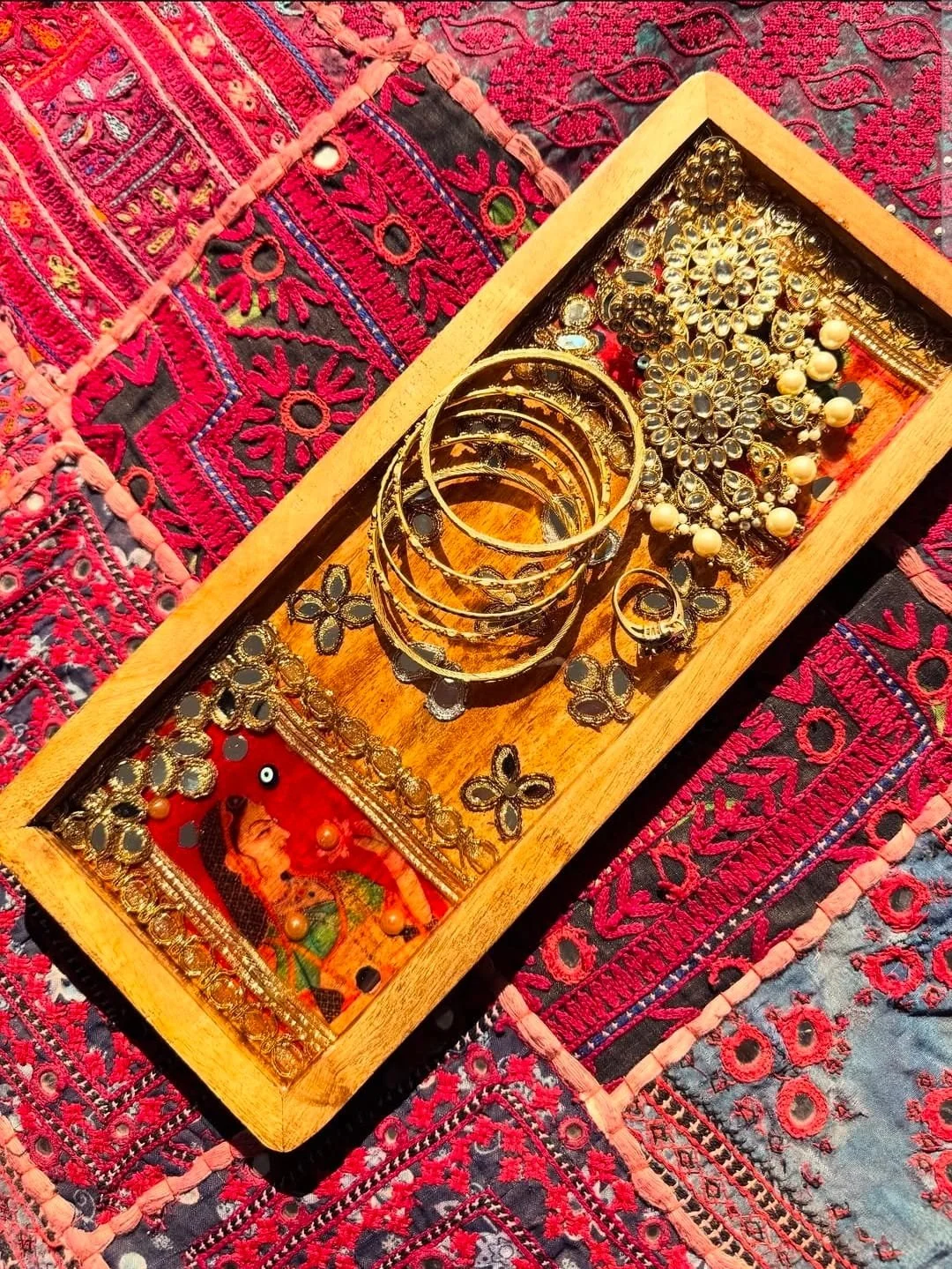 Display of gold earrings and bangles in a wooden tray, with ornate jewelry including earrings and bangles, on a colorful embroidered textile background.