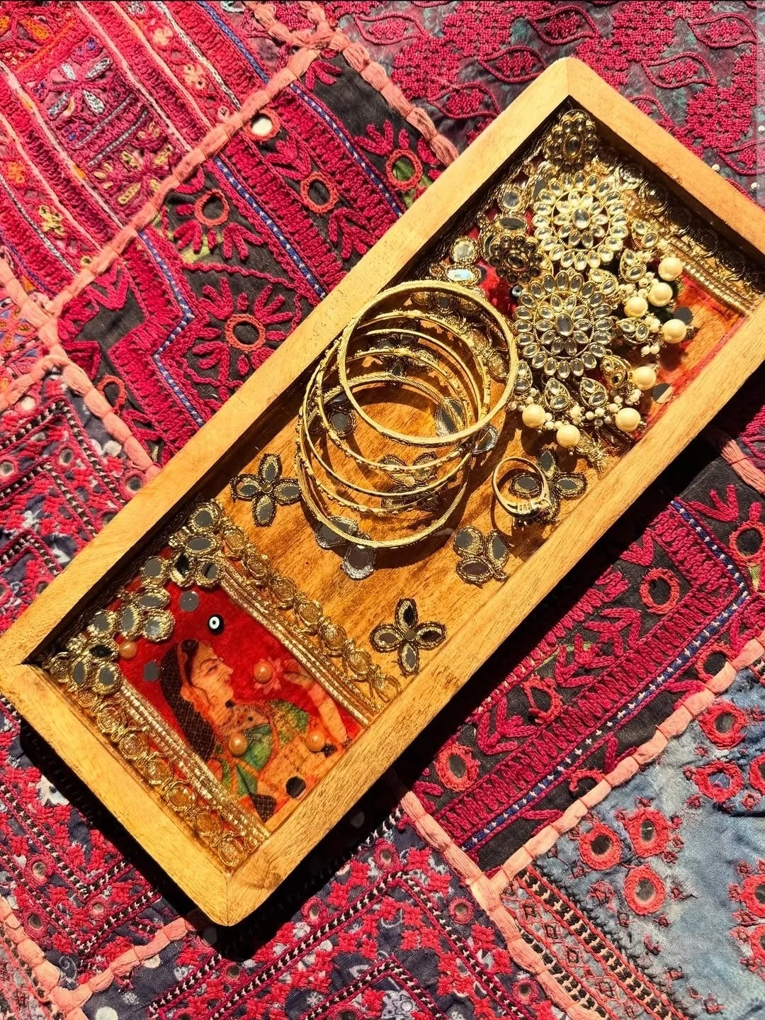 A wooden tray containing gold jewelry including bangles, earrings, and rings, placed on a colorful embroidered textile with red, pink, black, and green patterns.