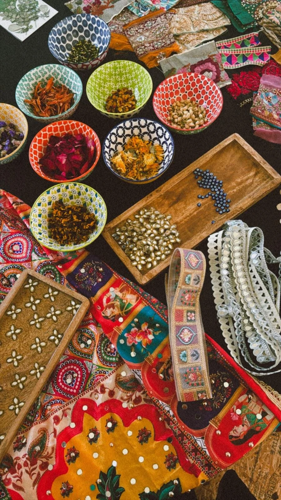 Colorful bowls with various dried herbs and flowers, embroidery and textile pieces, beads, and decorative fabric on a black background, indicating a craft or jewellery-making setup.