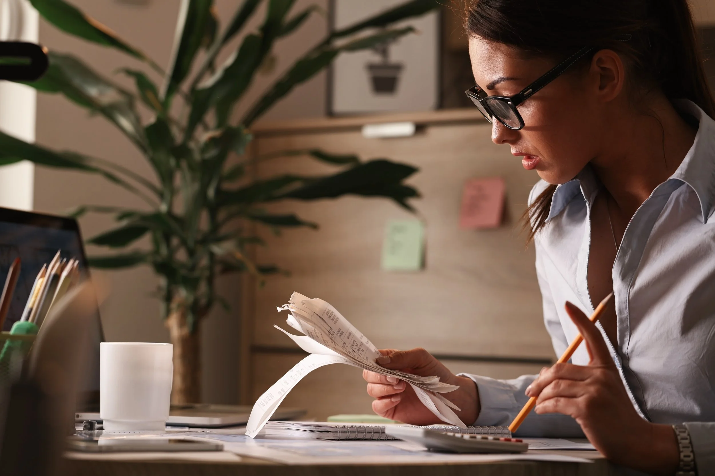 Woman with glasses sitting at desk, studying receipts and using calculator