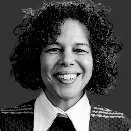Black and white portrait of a woman with short curly hair, wearing a collared shirt, a cardigan, and layered necklaces, smiling softly.